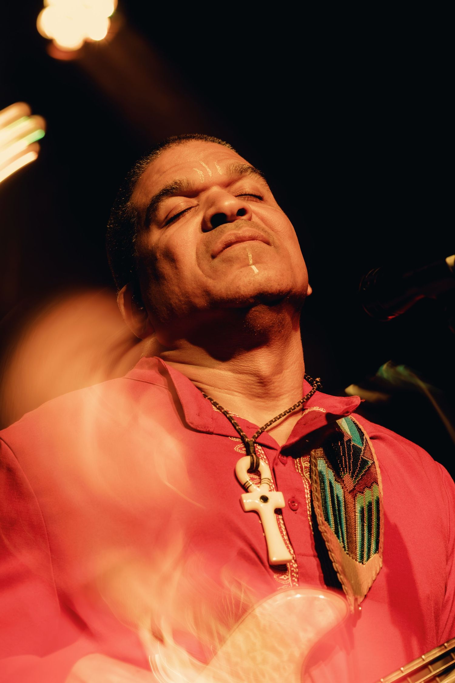 Man in pink shirt with cross necklace plays guitar on stage under warm lights.