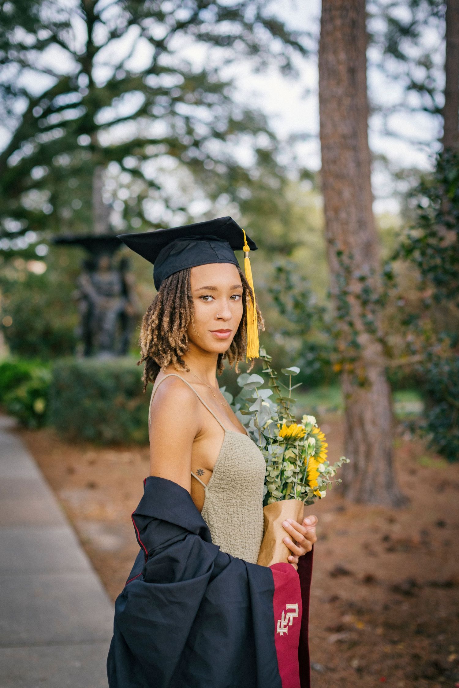 Graduate in graduation cap and gown stands outdoors with trees and flowers while holding diploma and yellow bouquet.