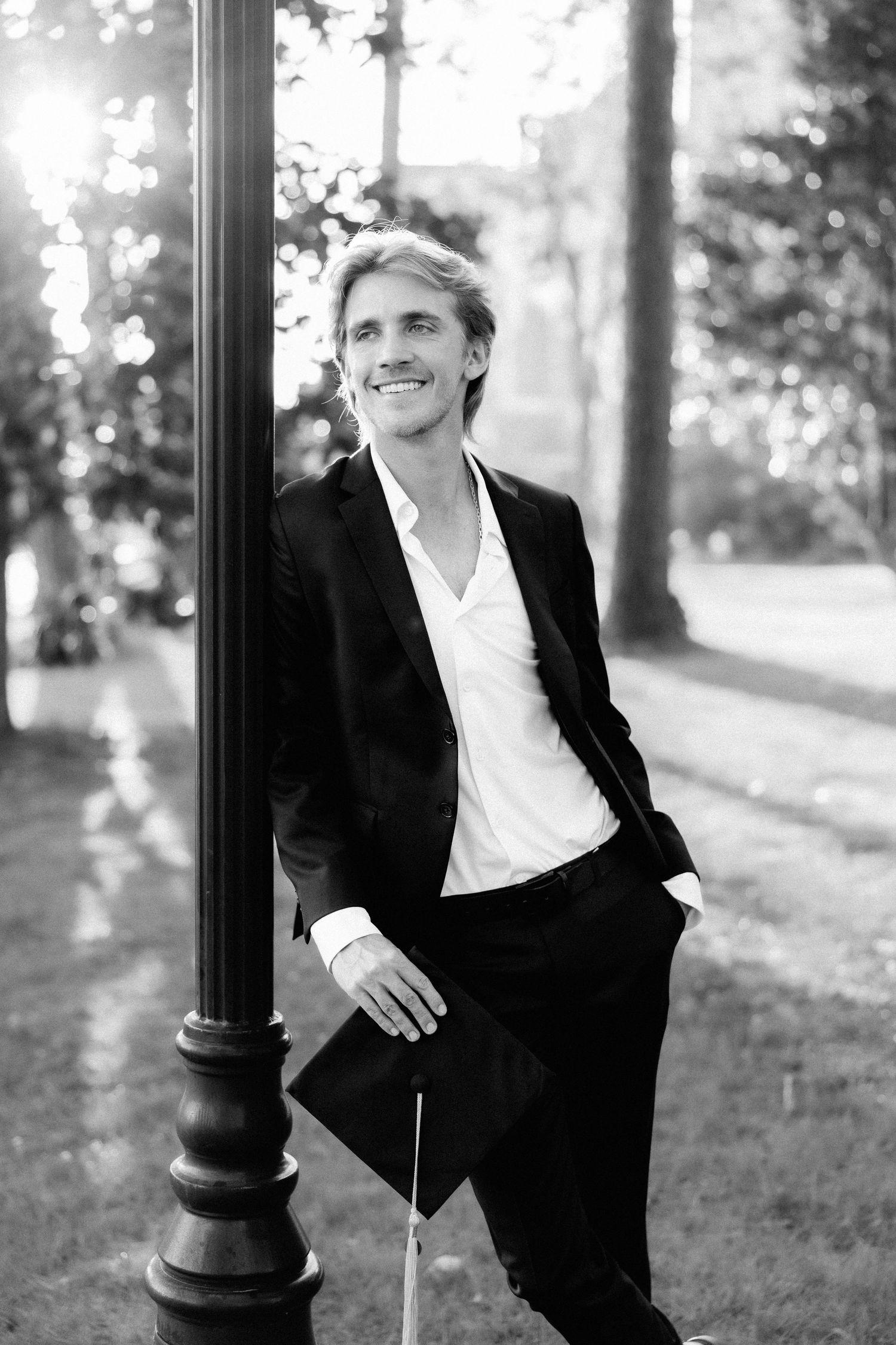 Smiling man in black suit leaning against lamp post in park, black and white portrait photography.