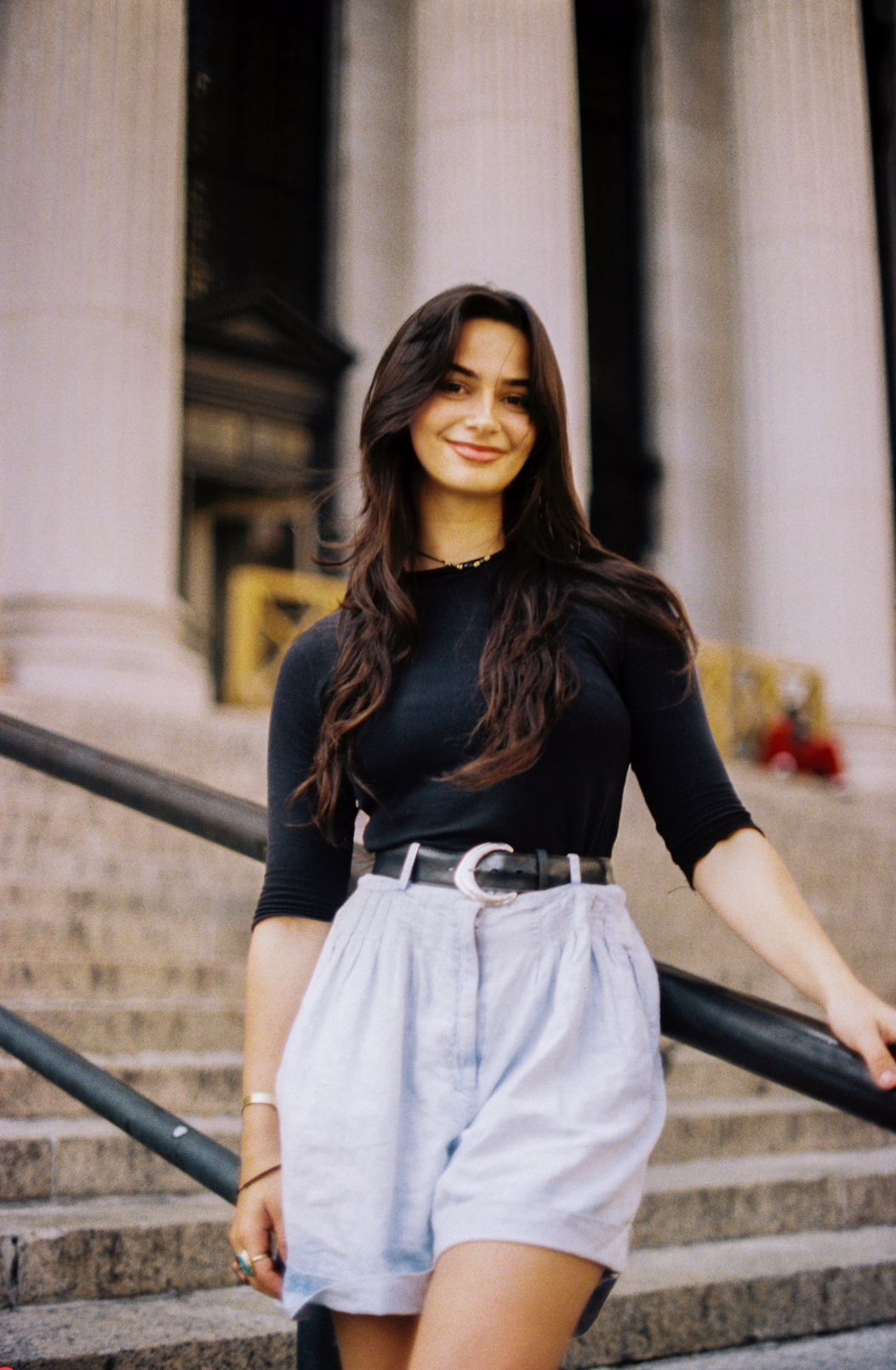 A woman in a black top and high-waisted white shorts stands smiling on stone steps near classical columns.