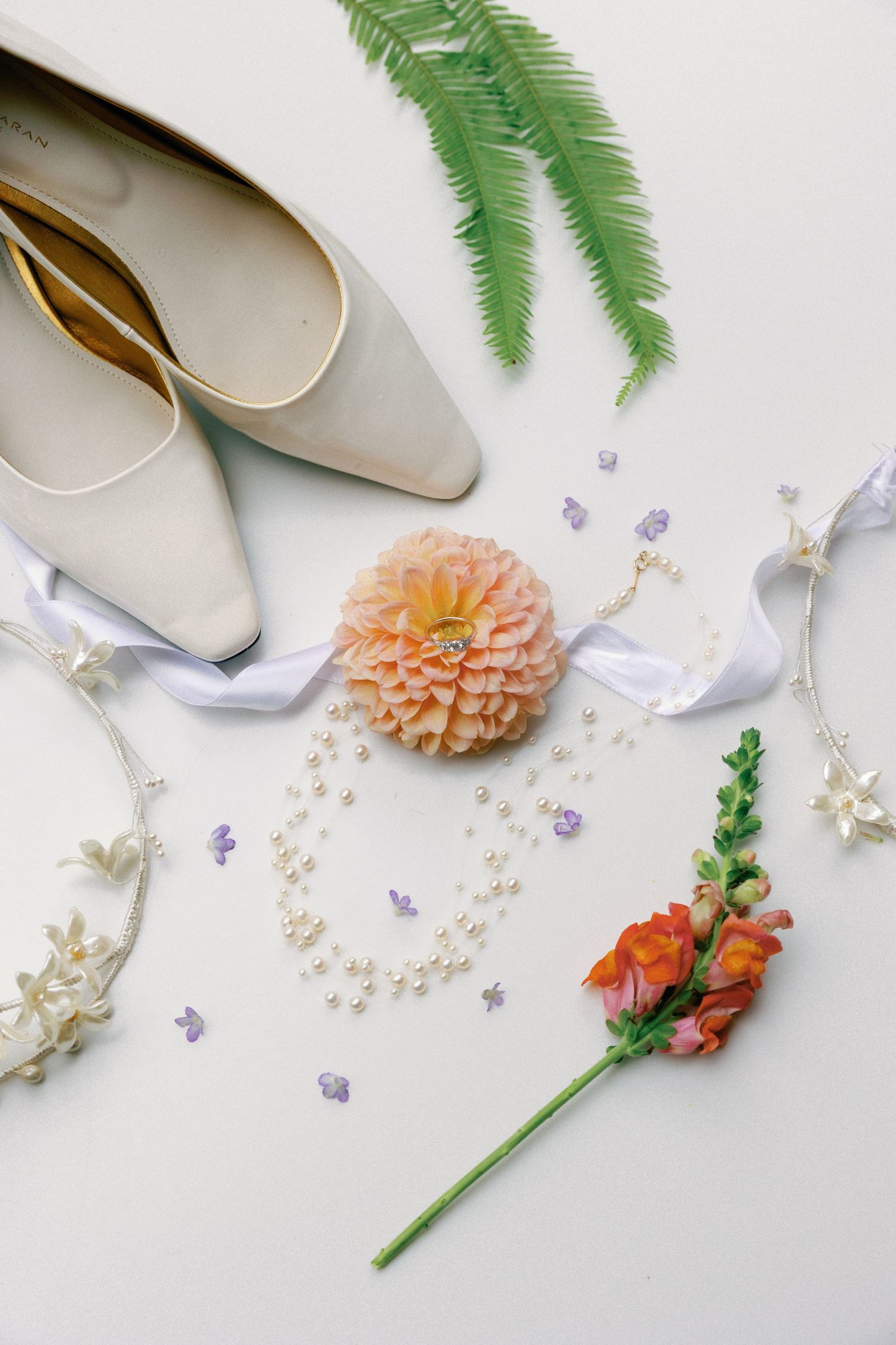 Cream pointed-toe flats, pearl strands, a peach dahlia, orange snapdragon and fern leaves arranged as bridal flat lay accessories.