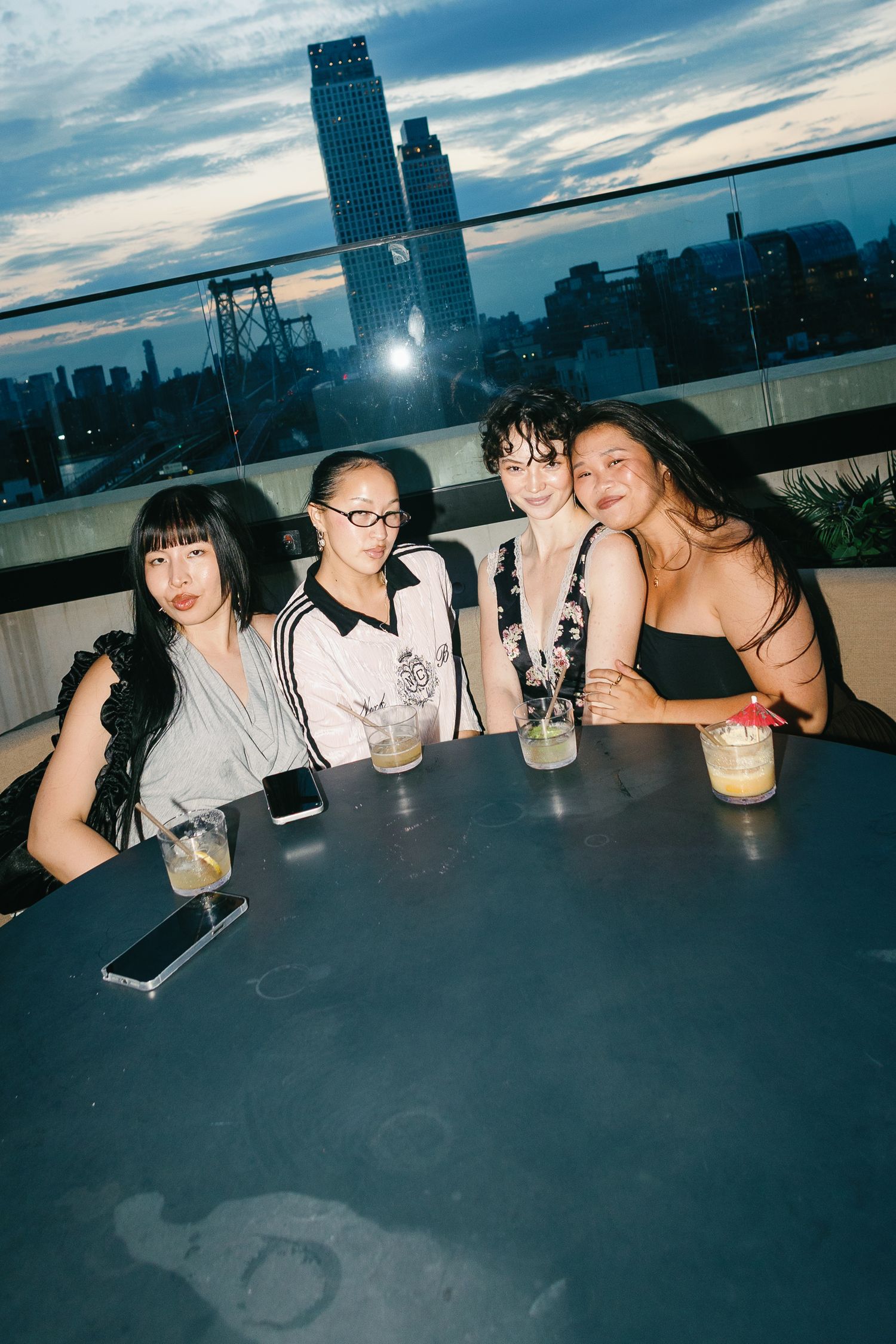 Four young women smiling together at a rooftop bar table with cocktails, city skyline at dusk in background.