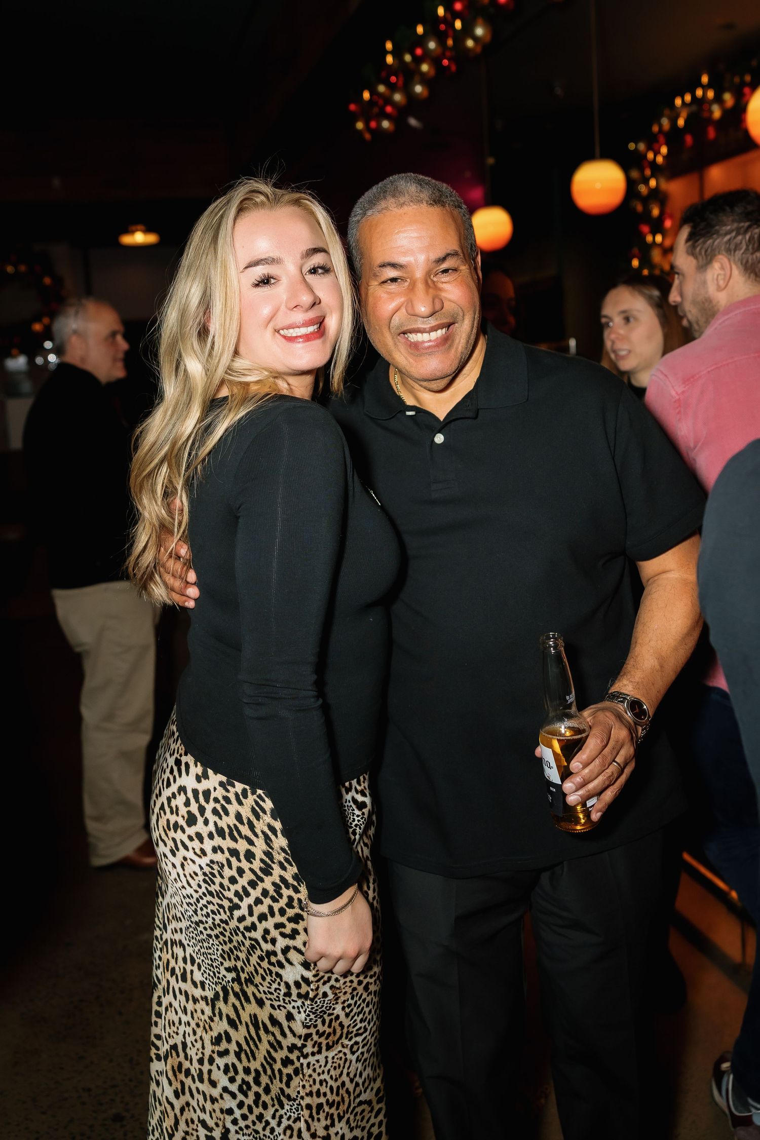 A smiling blonde woman in leopard print skirt and man in black polo holding a beer pose together at a social event.