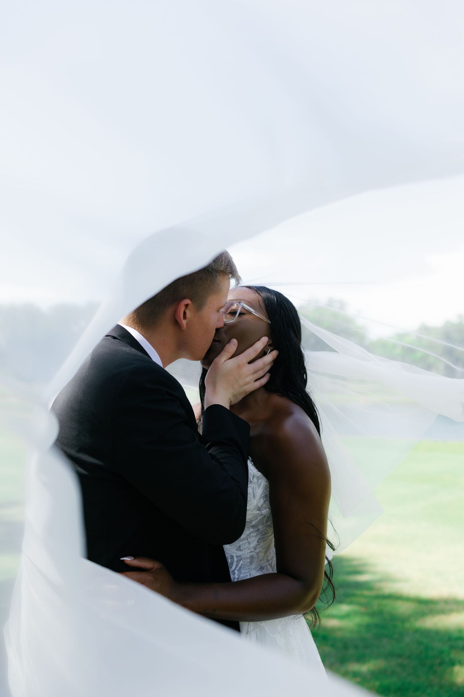 A bride and groom share a romantic kiss outdoors, wrapped in a flowing white veil on their wedding day.