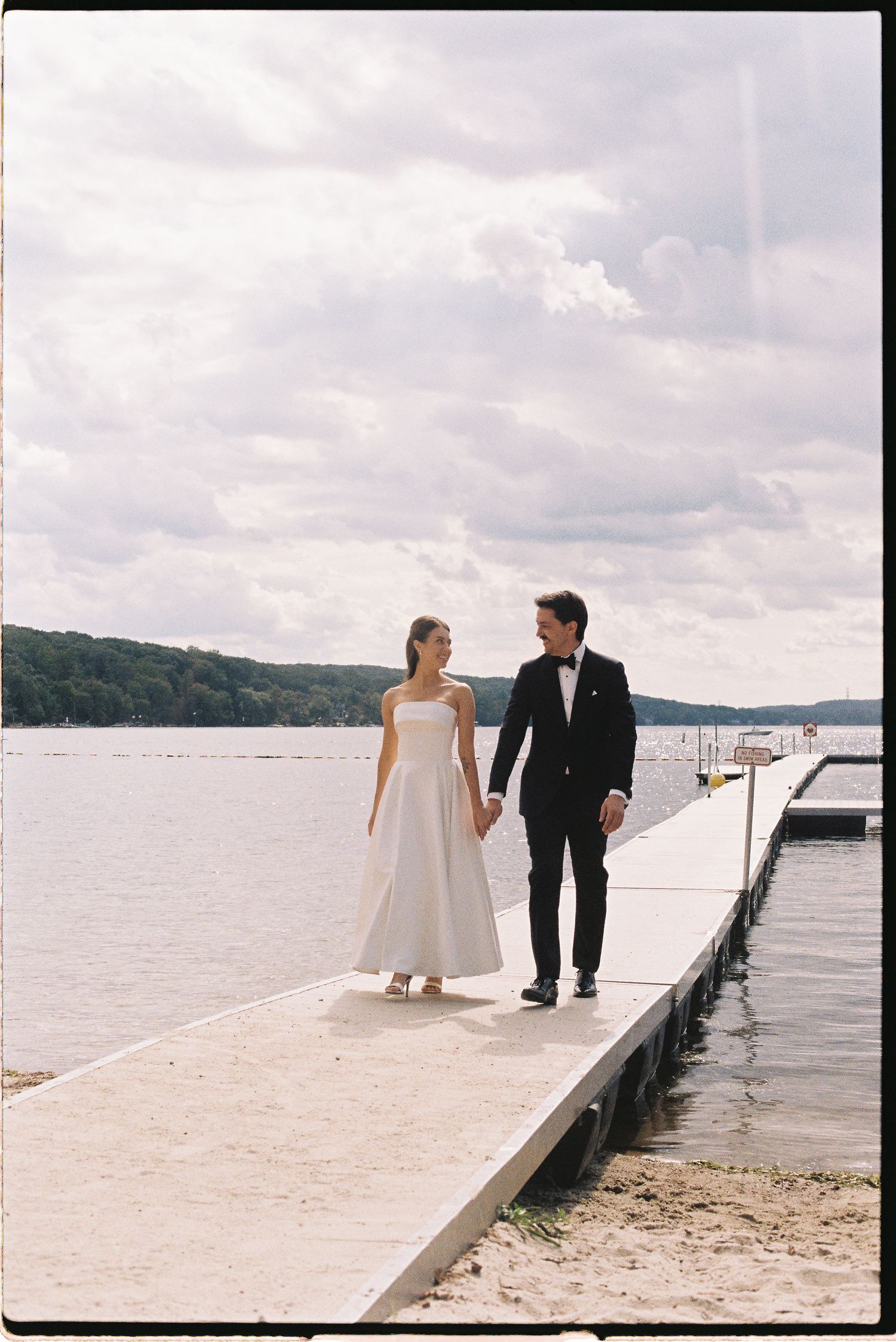 Bride and groom hold hands walking on a lakeside dock, wearing wedding attire with cloudy skies and trees in background.