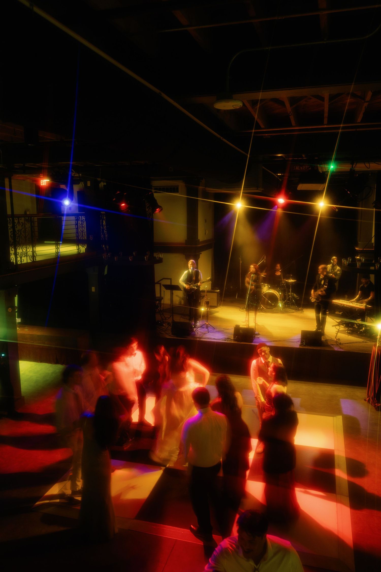 Live band performing on stage with colorful lights as guests dance on the floor at an indoor evening event.