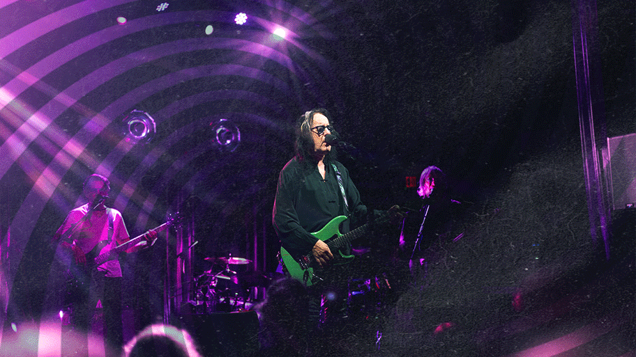 Concert performers on stage illuminated by vibrant purple lighting effects during a live music performance.