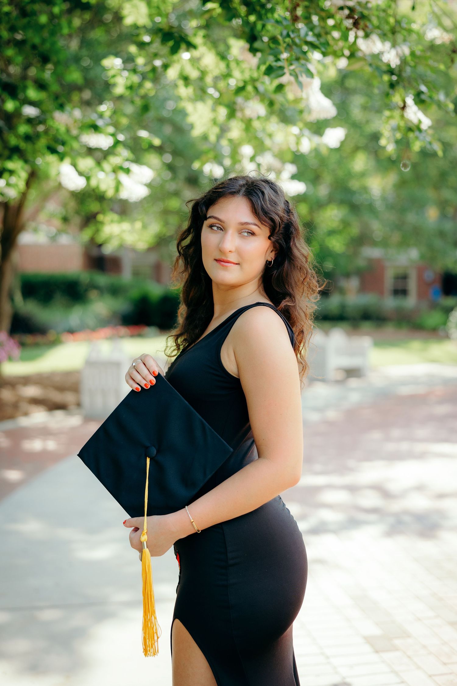 A graduate in a black dress poses outdoors on a sunny day with trees and dappled light in the background.