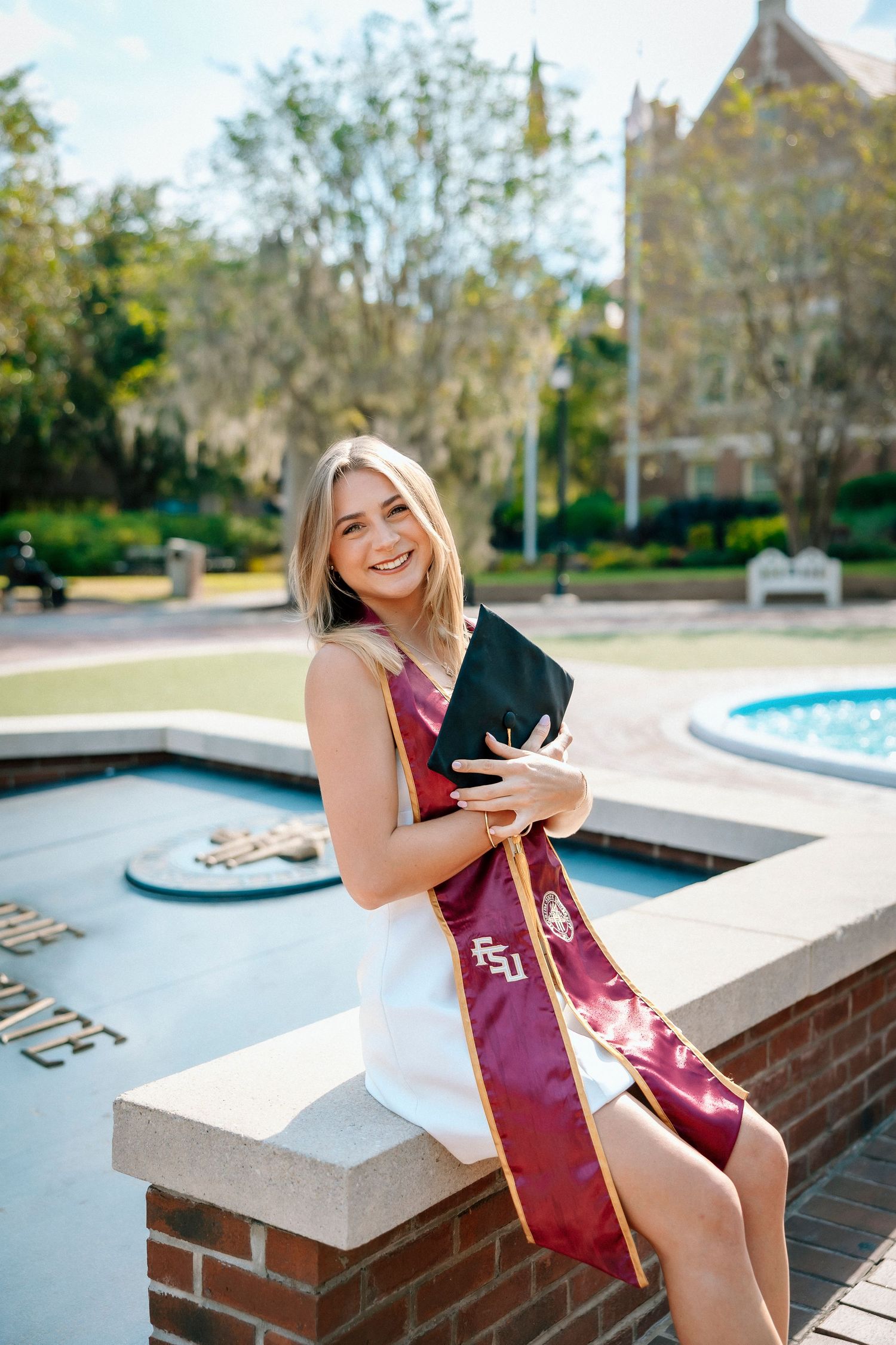 Graduate in maroon and gold sash poses by campus fountain for outdoor graduation photos on sunny day.