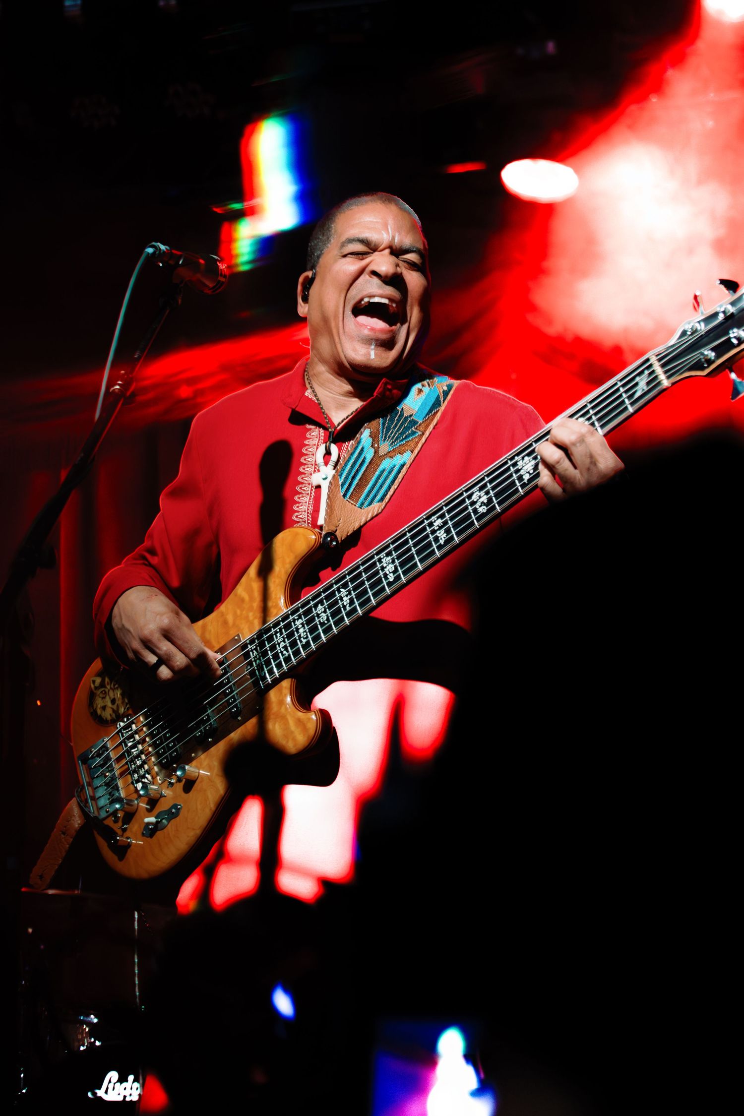 Energetic bassist in red shirt performs on stage, playing electric guitar under dramatic red stage lighting.