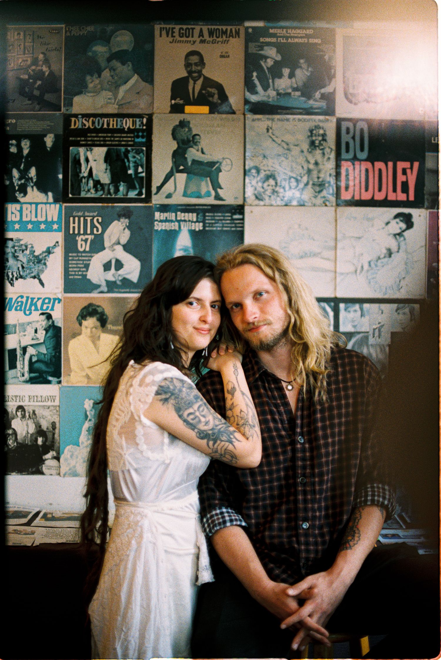 A tattooed woman in a white dress embraces a bearded man with long blond hair in front of a wall of vintage vinyl record covers.
