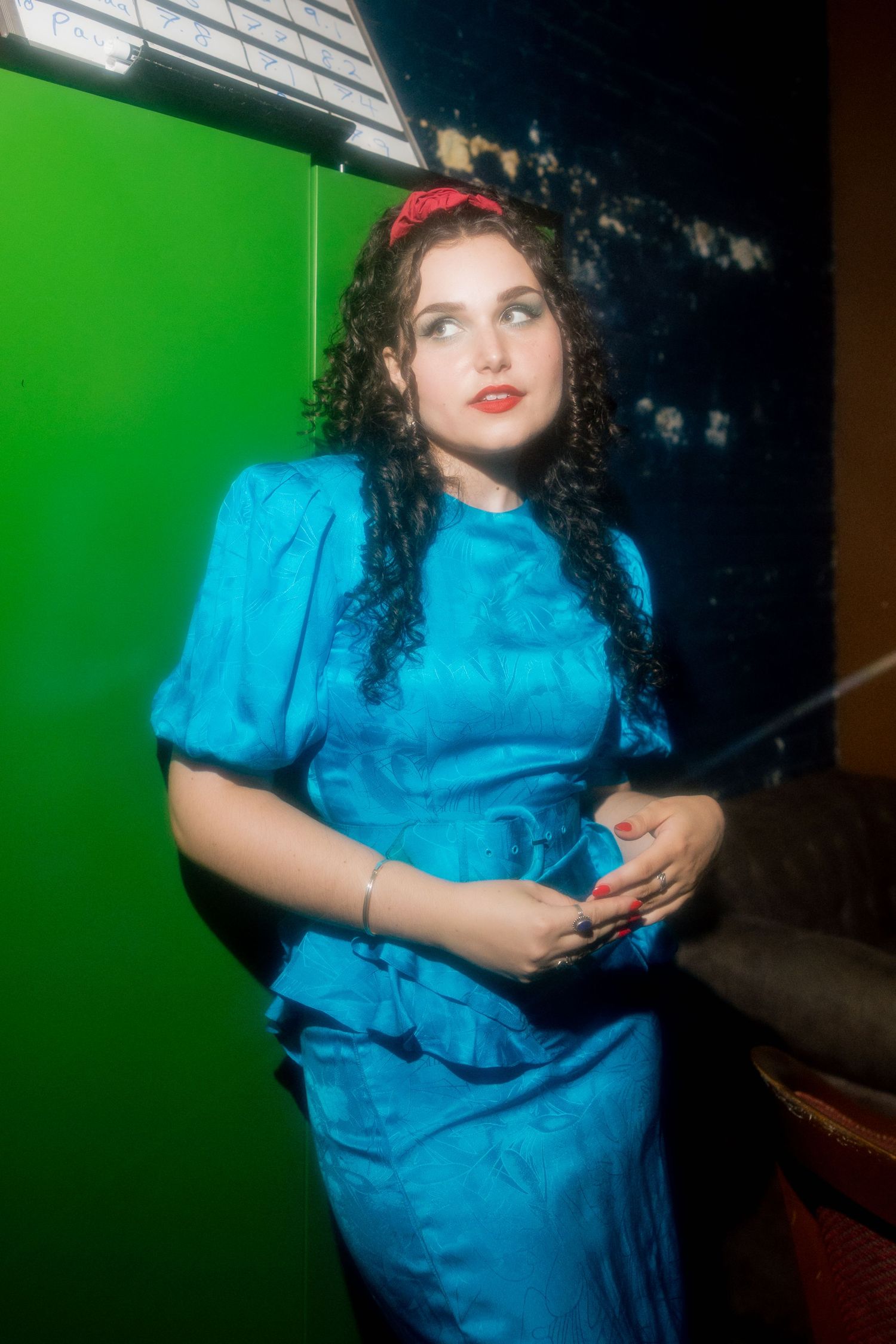 Woman in blue satin dress with red headband and curly hair poses near green screen backdrop.