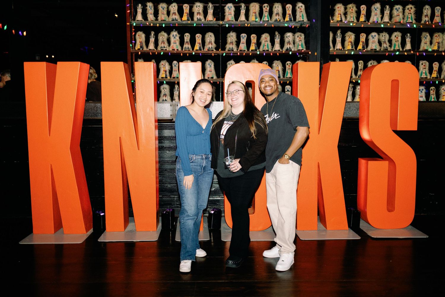 Three people posing in front of large orange KNICKS letters sign at a sports bar event.