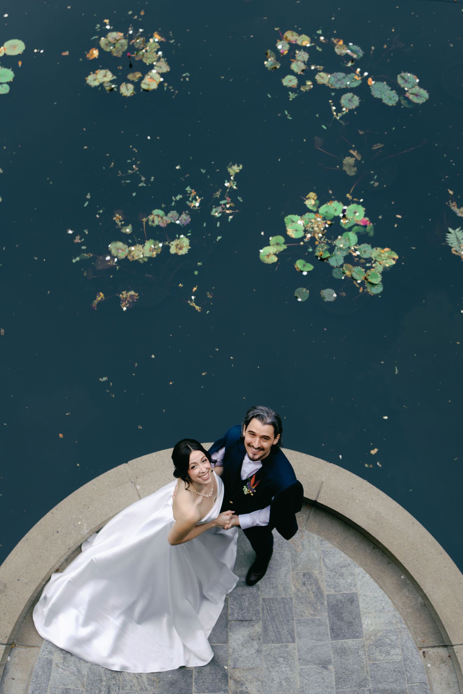 Aerial view of a smiling bride and groom embracing on a curved stone ledge beside a dark pond dotted with water lily pads.