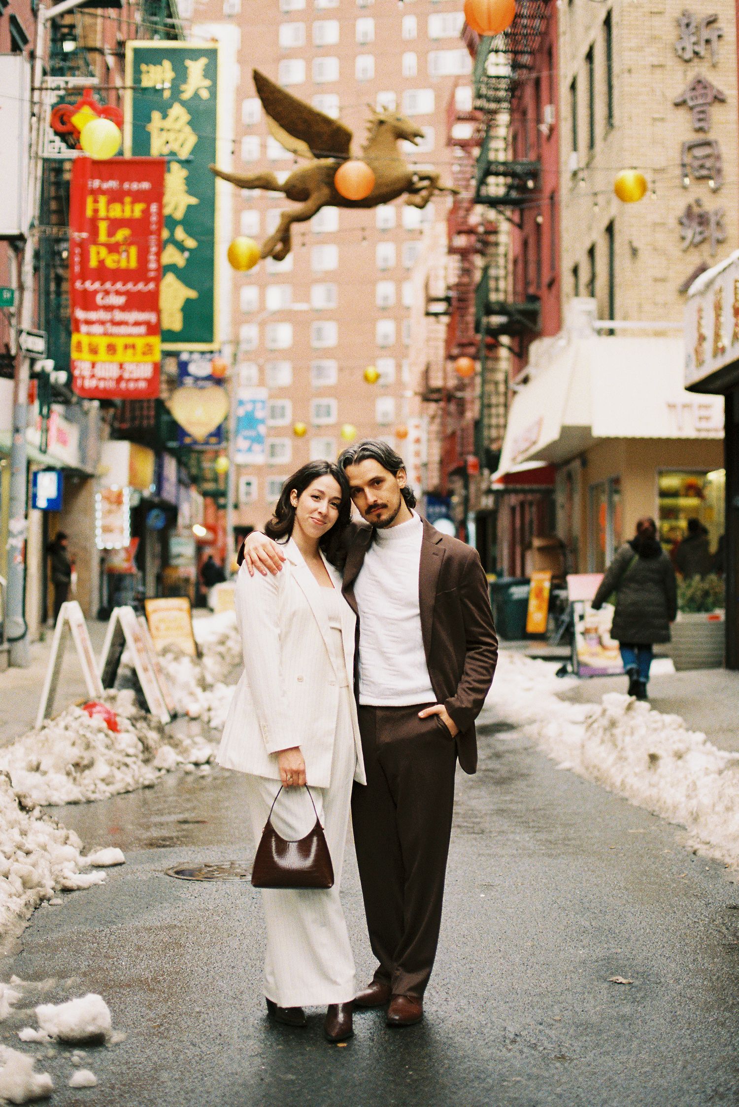 A couple walks down a snowy street in Chinatown, New York City with lanterns and decorations visible.