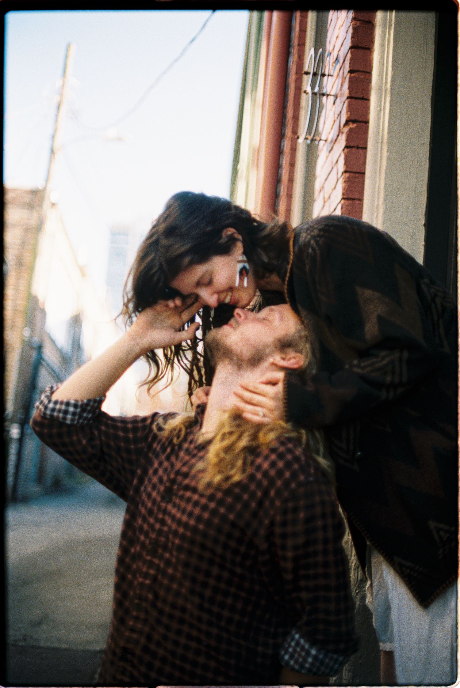 A smiling woman with long dark hair leans down to kiss a bearded man on a brick-lined city street in warm, vintage-style light.