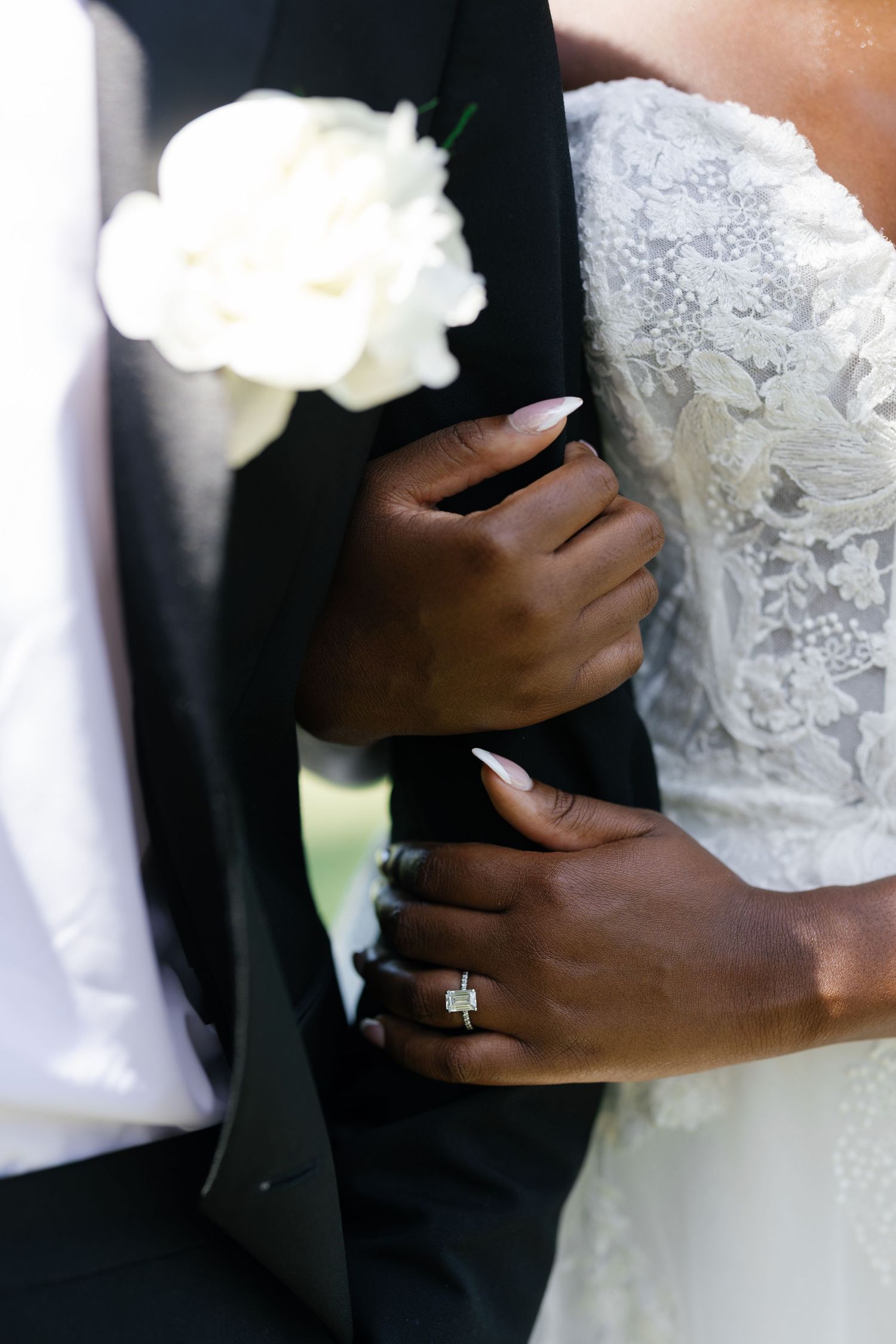 Close-up of bride's hands with manicured nails and diamond ring embracing groom's black tuxedo jacket.