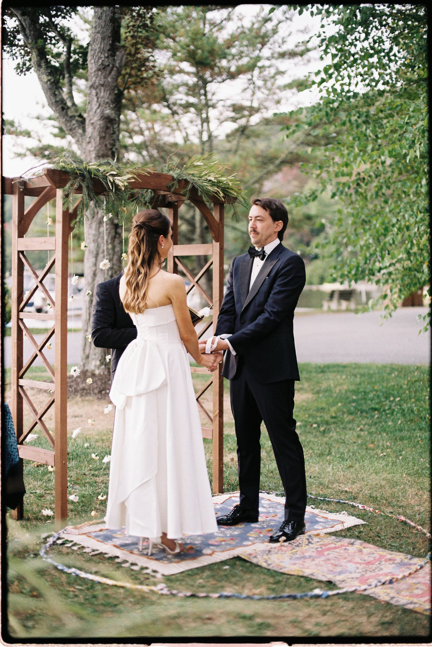 Bride and groom hold hands at outdoor wedding altar under wooden arbor with greenery on layered rugs.