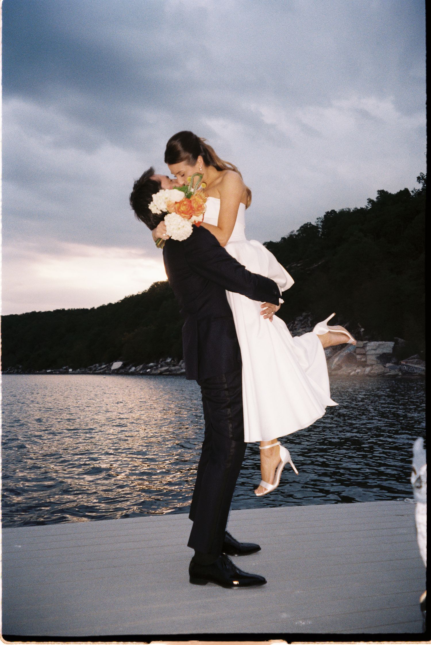 Groom lifts bride in white strapless gown on lakeside dock at sunset wedding.