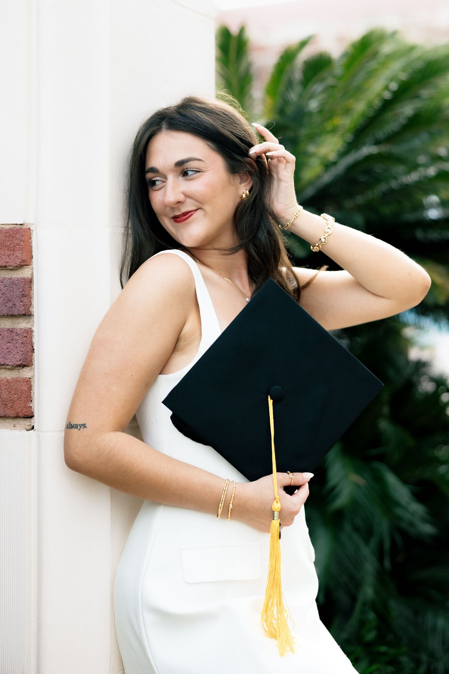 Young woman in white dress holding black graduation cap with gold tassel, smiling outdoors near greenery.