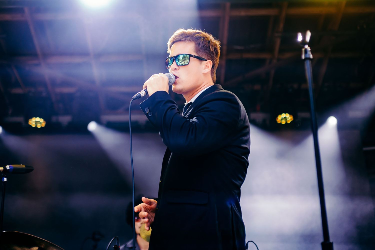 A performer in sunglasses and dark clothing sings into a microphone on stage with dramatic lighting in the background.