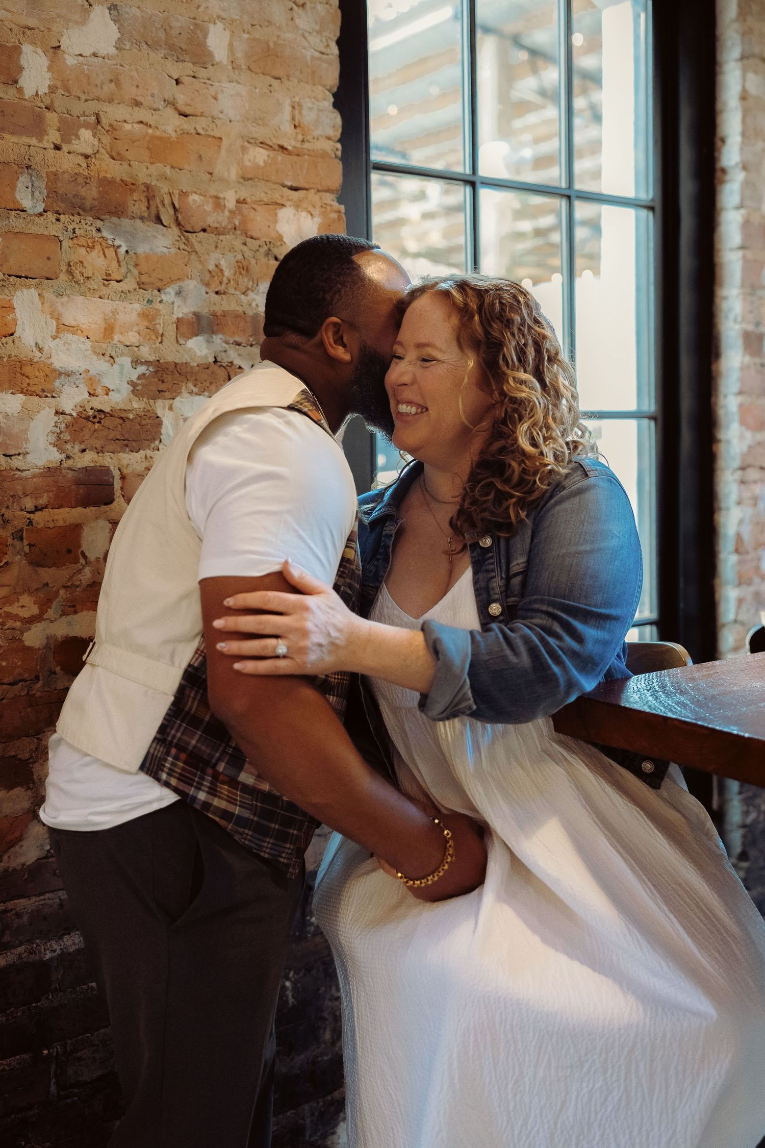 A romantic couple shares an intimate moment together by a sunlit window in a rustic brick-walled space.