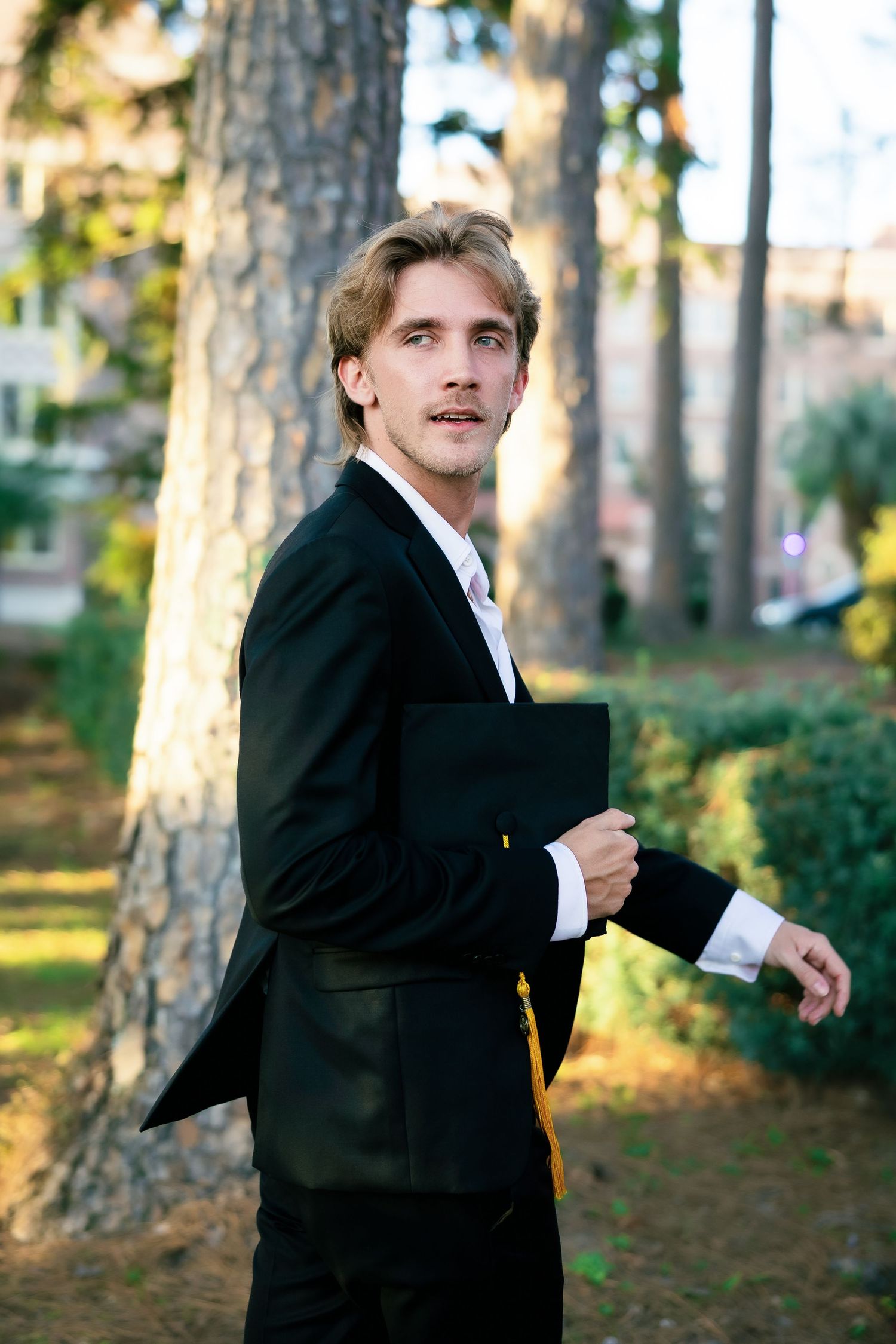 Young man in black suit holding a folder outdoors among tall trees in a sunlit park setting.