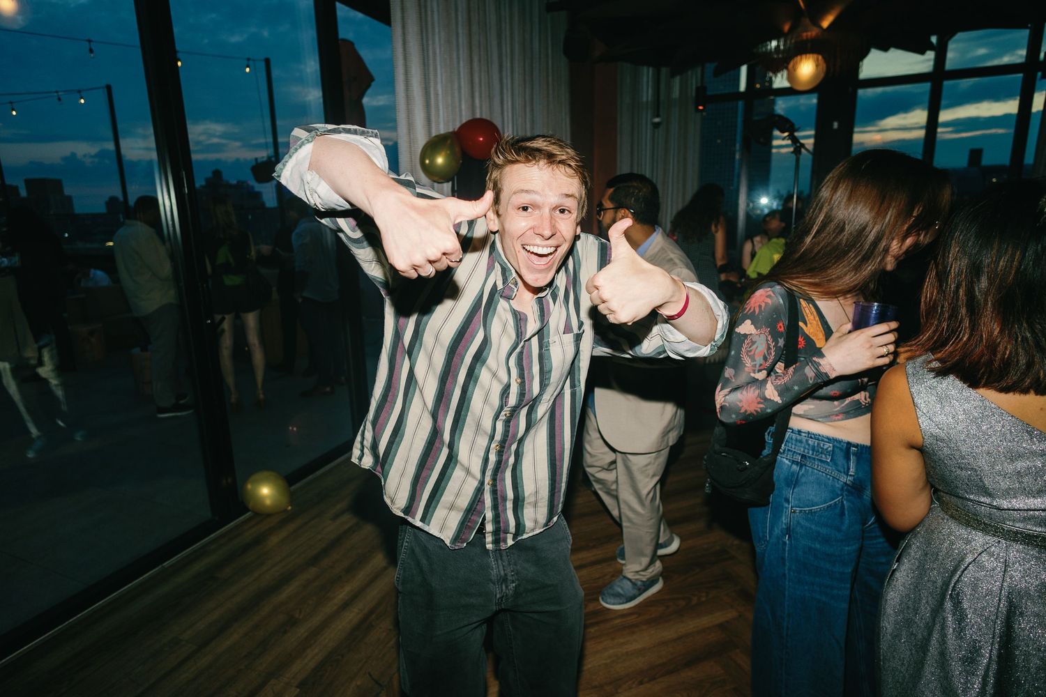 Young man in striped shirt giving thumbs up while dancing at a party venue with large windows.