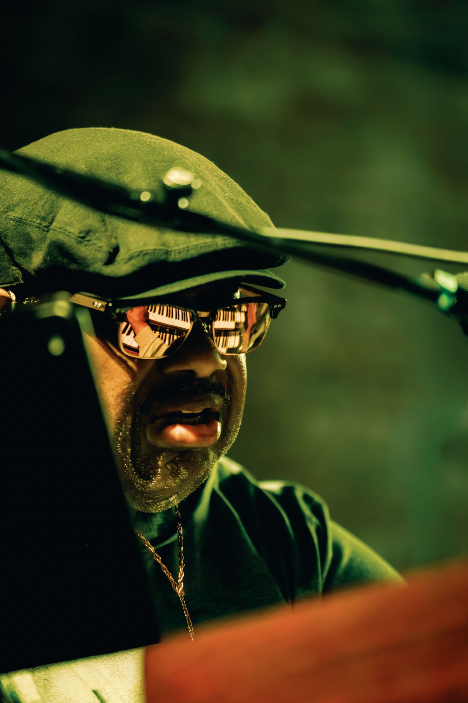 A bearded man wearing a green flat cap and reflective sunglasses, with a piano reflected in the lenses.