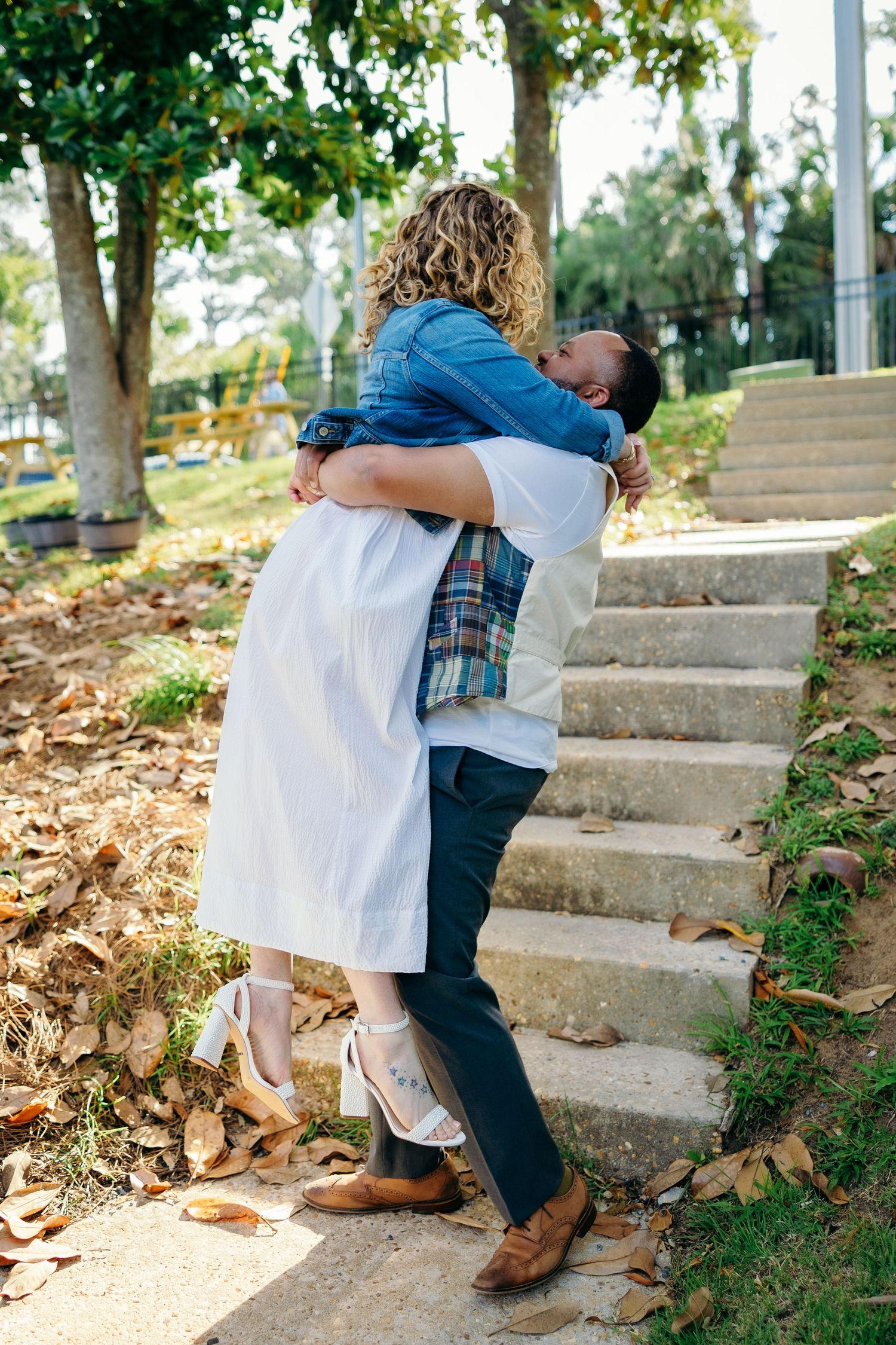 Two people embrace in a tender moment on outdoor stone steps surrounded by greenery and fallen leaves.
