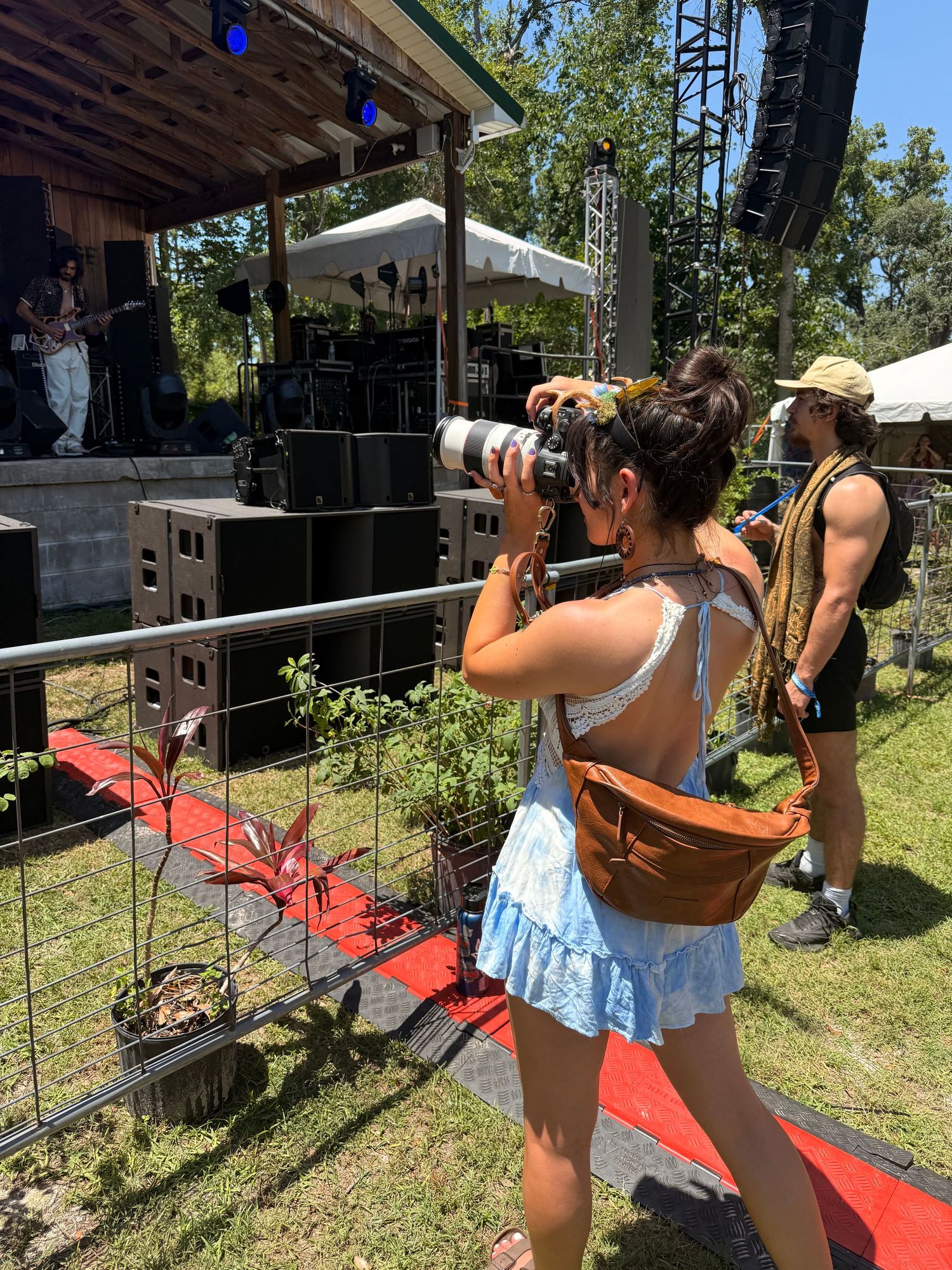 A photographer with a professional camera stands along a red-roped barrier near an outdoor stage during a sunny event.