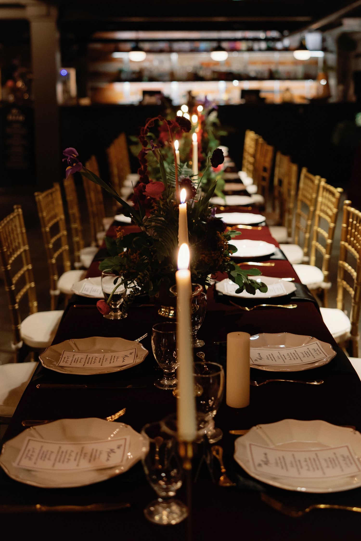 Elegant candlelit wedding reception table with gold chairs, dark linens, floral centerpieces, and white menu cards in moody lighting.