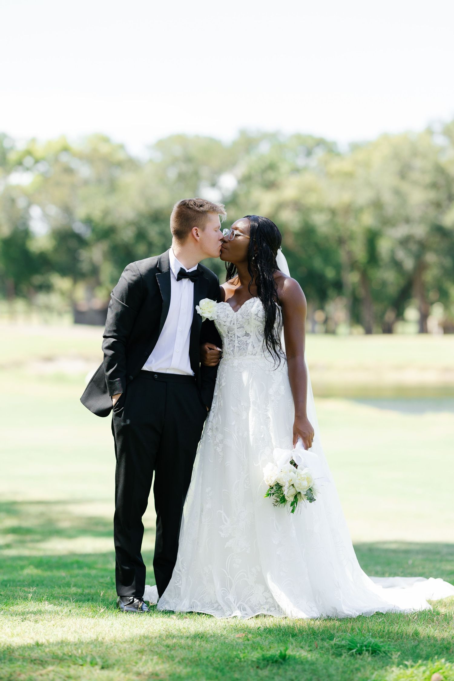 Interracial couple sharing a kiss outdoors, bride in lace gown holding white bouquet, groom in tuxedo.