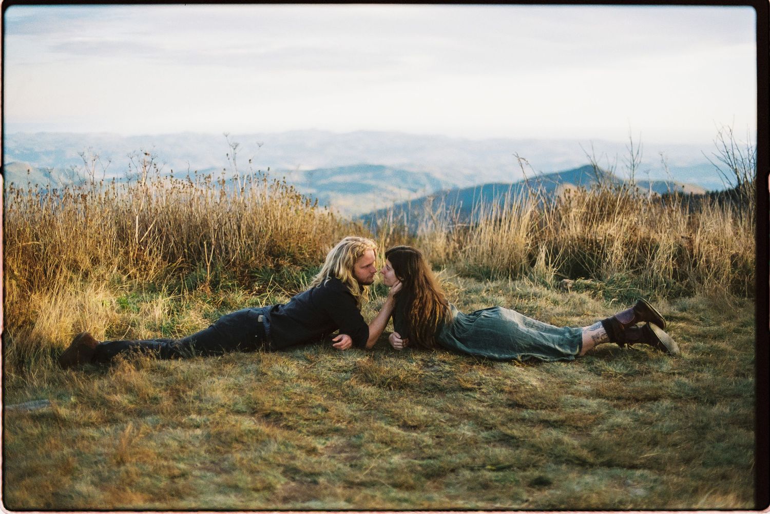 Two people lie face to face on a grassy mountaintop with golden grass and hazy blue mountain ridges stretching into the distance.