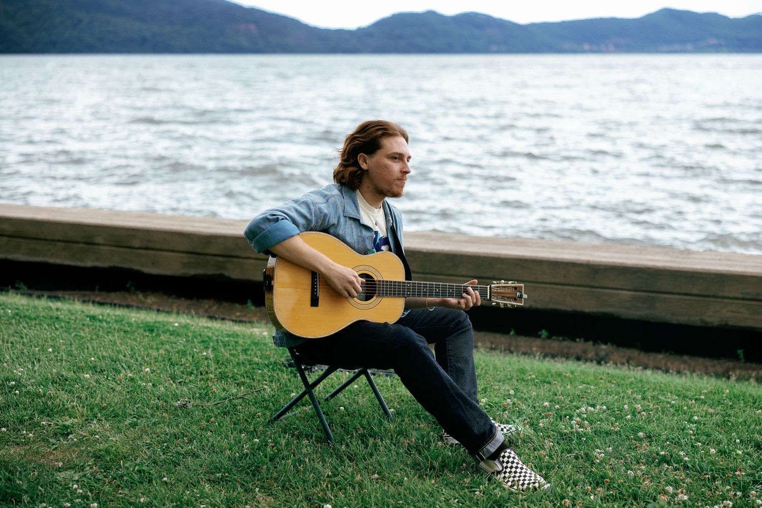 Young man with long hair playing acoustic guitar on grass by a scenic lake with mountains in background.