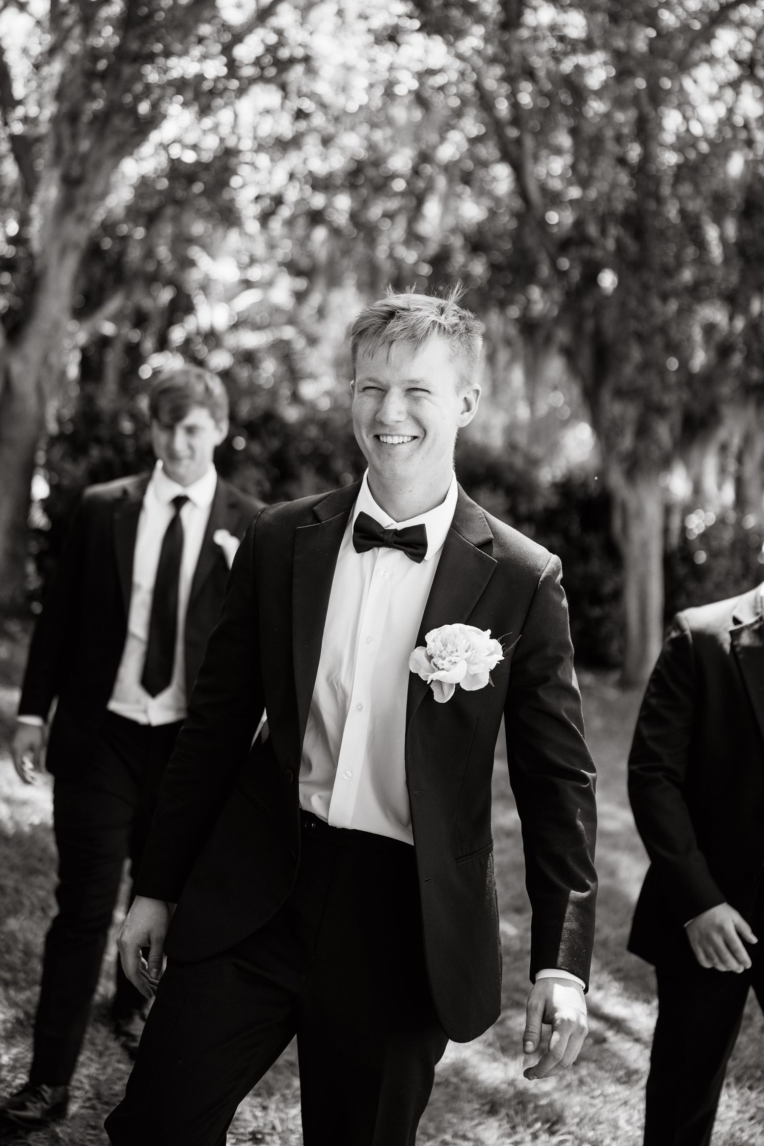Smiling groom in black tuxedo with boutonniere walks with groomsmen outdoors in black and white photo.