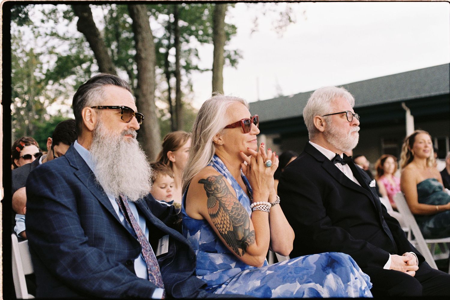 Three adults in formal attire sit outdoors at a wedding ceremony, smiling and clapping.