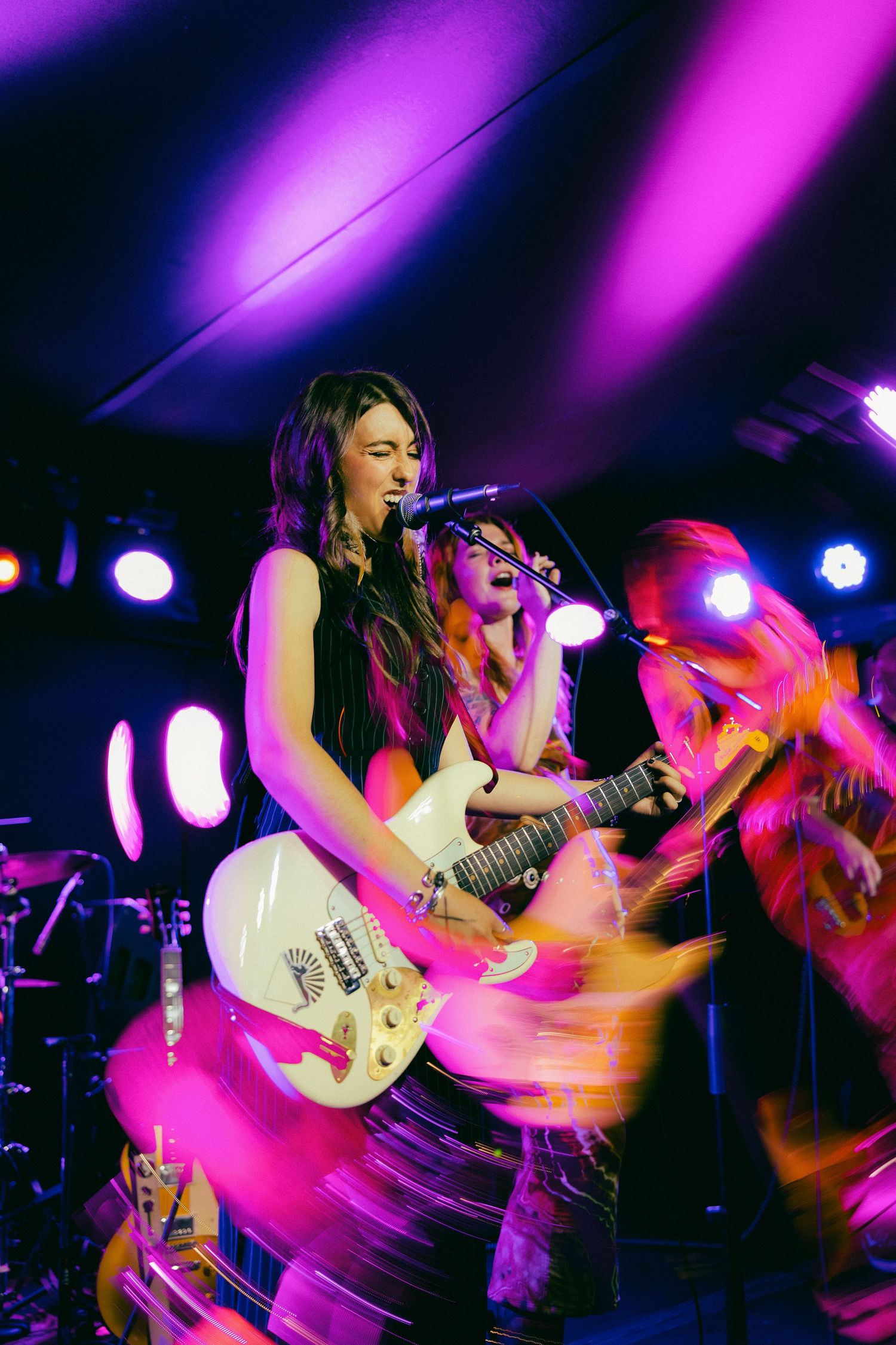 Musicians perform energetically on stage under vibrant purple lighting with electric guitars and dramatic motion blur effects.
