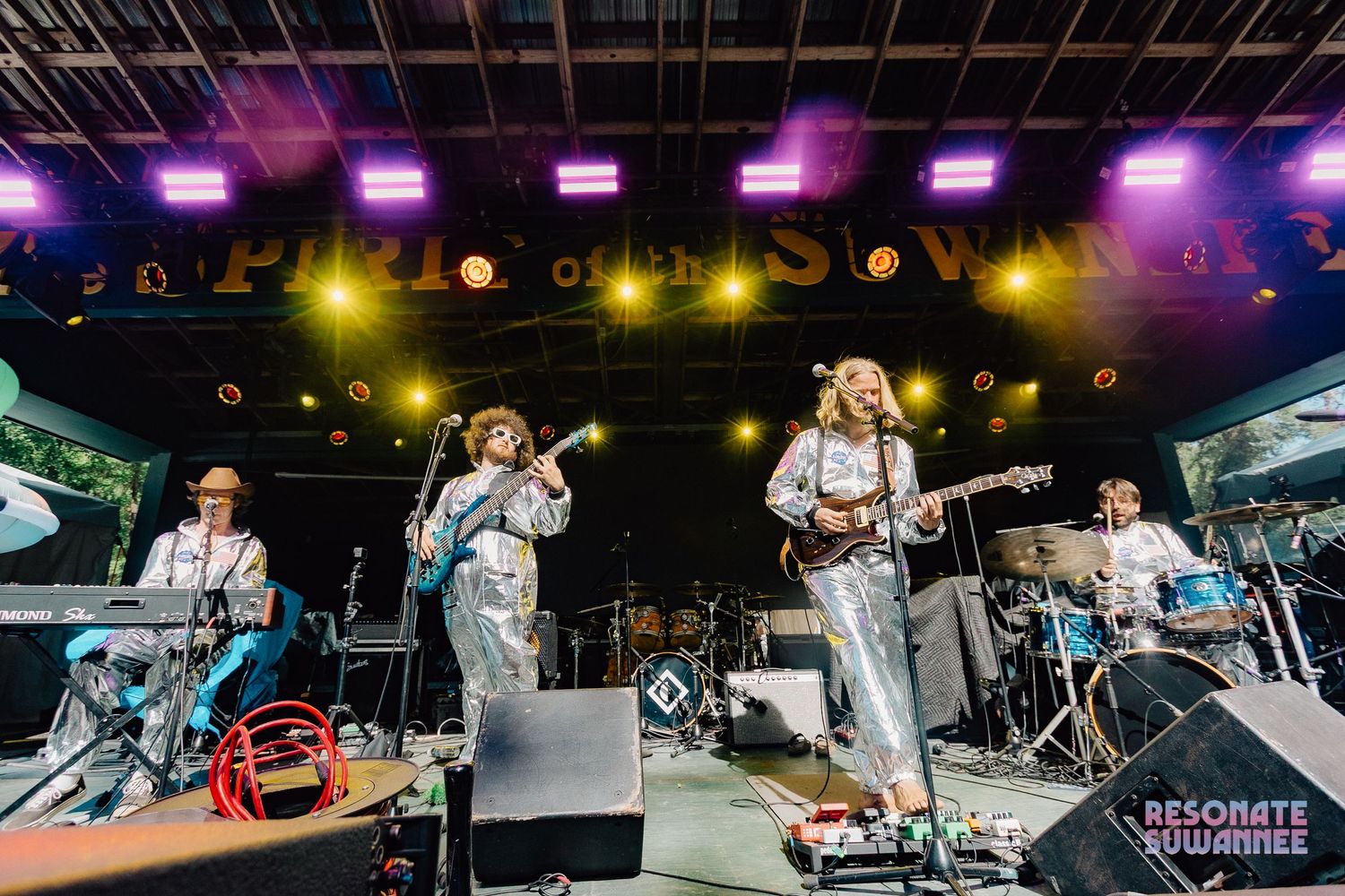 A live band performs on stage with vibrant purple and yellow lighting during an outdoor concert performance.