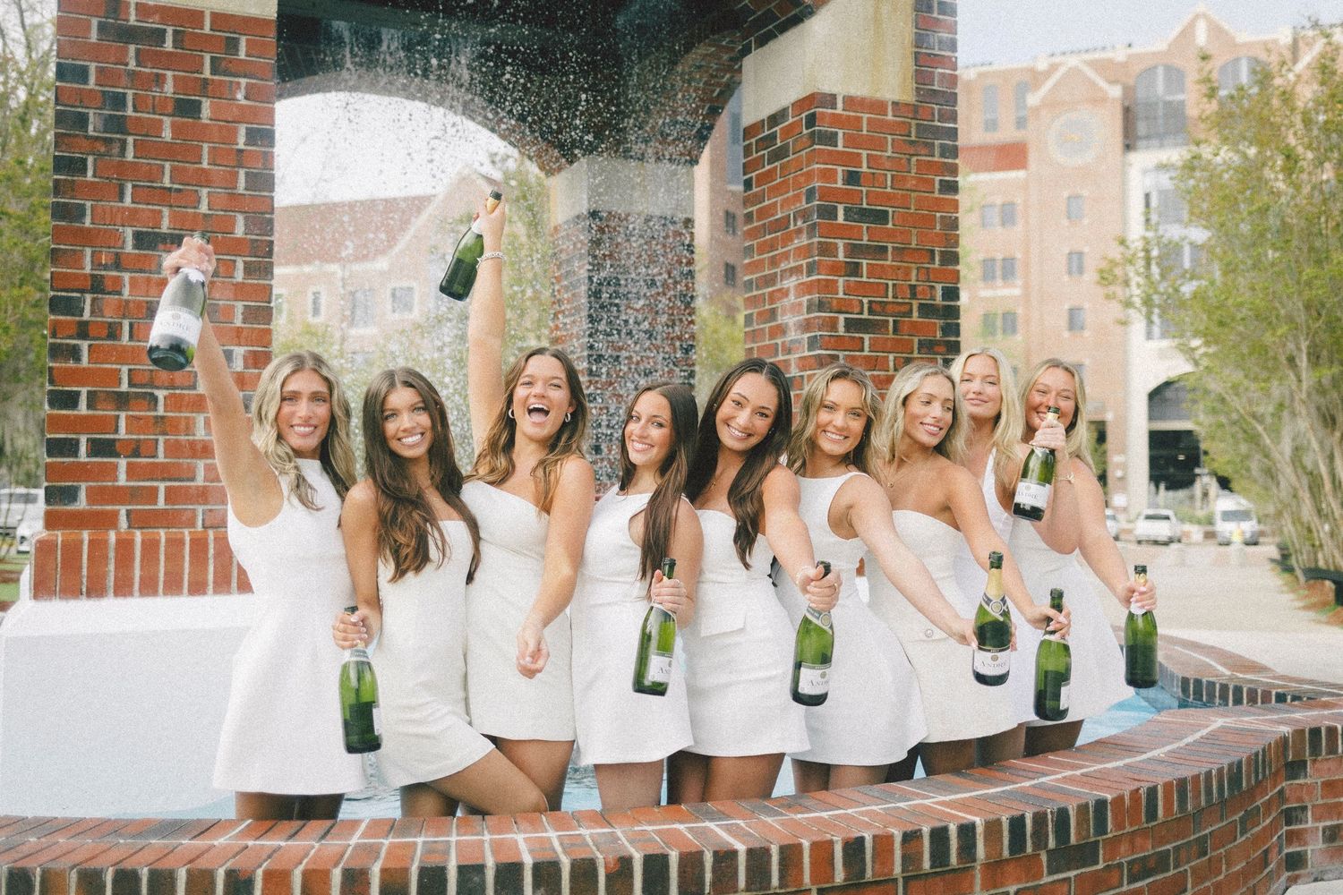 A group in white dresses sits on a brick wall while popping champagne bottles in celebration.