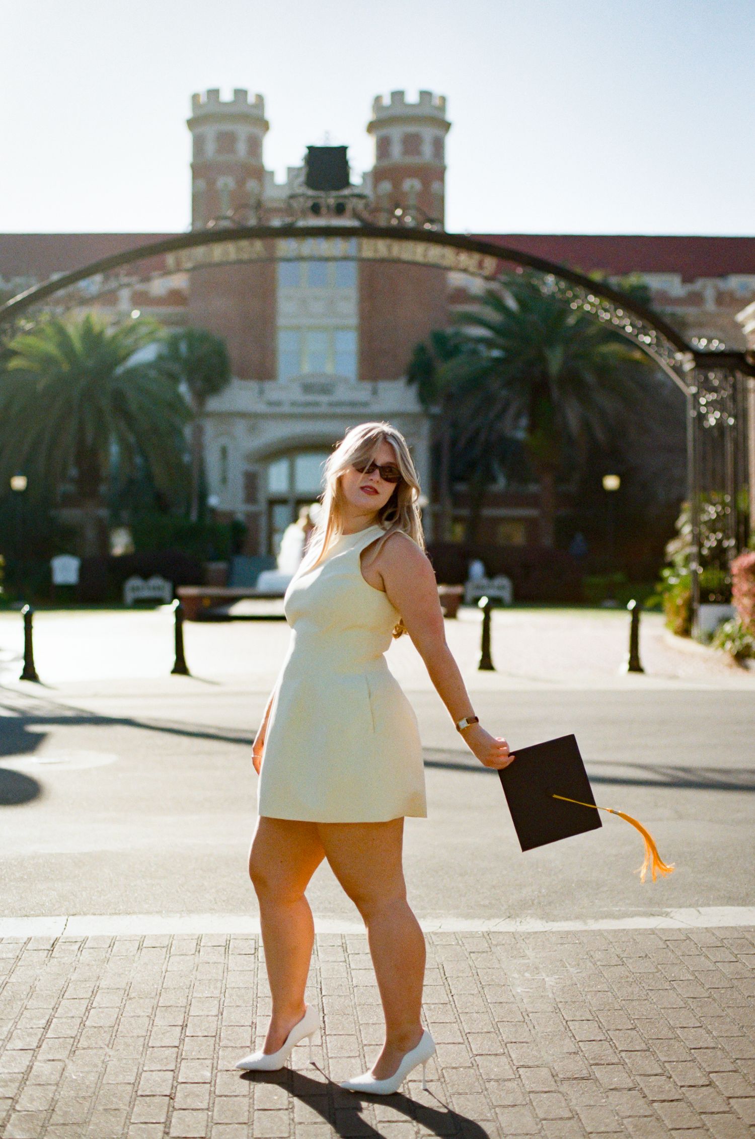 A woman in a white dress holds a graduation cap with gold tassel in front of a brick university entrance gate.