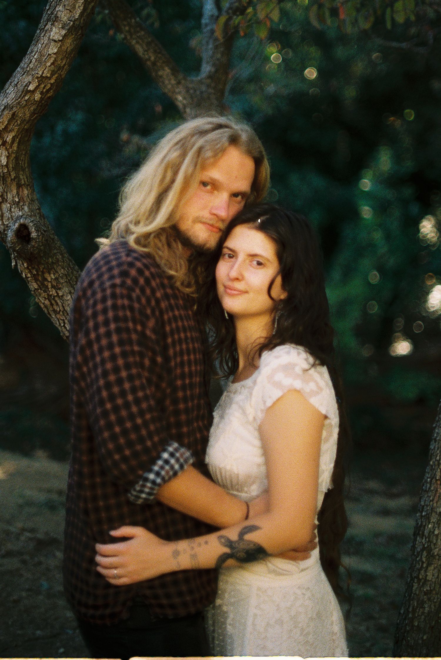 A couple embraces beneath a tree in soft natural light, he in a plaid flannel shirt and she in a white lace dress with a wrist tattoo.