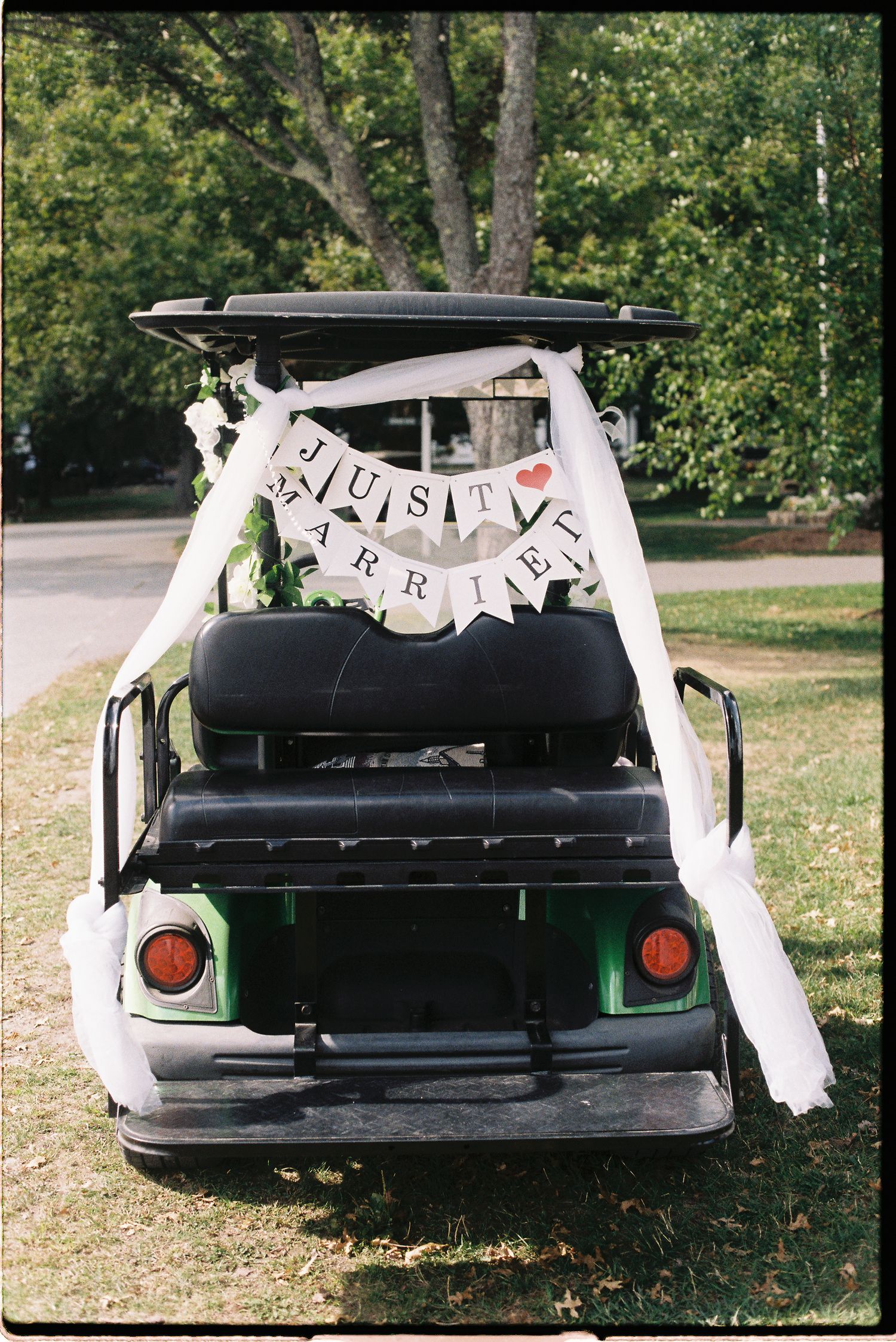 Green golf cart decorated with white ribbon and a Just Married banner on grass at an outdoor wedding.