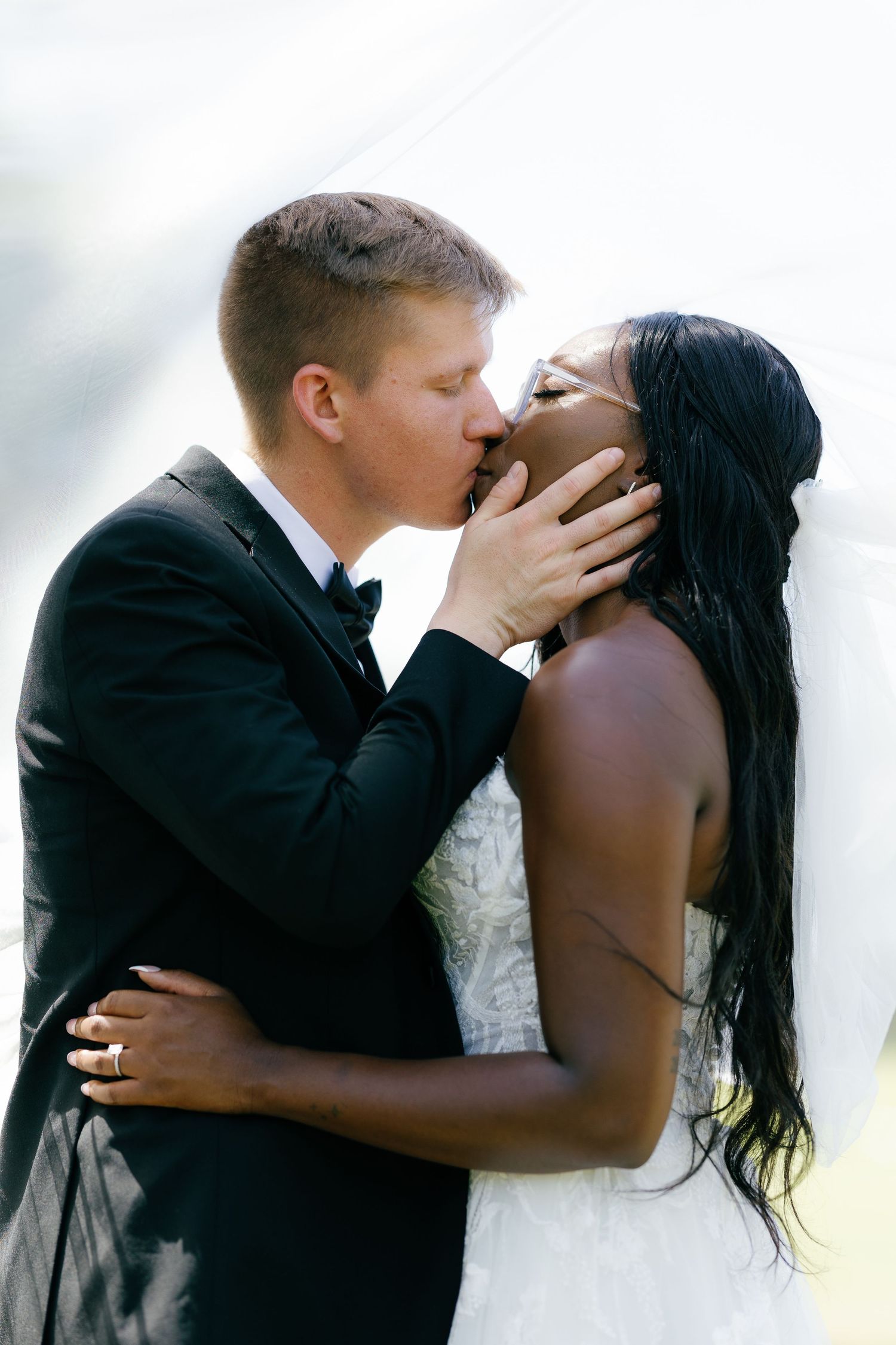 Interracial bride and groom sharing a romantic first kiss at their outdoor wedding ceremony.