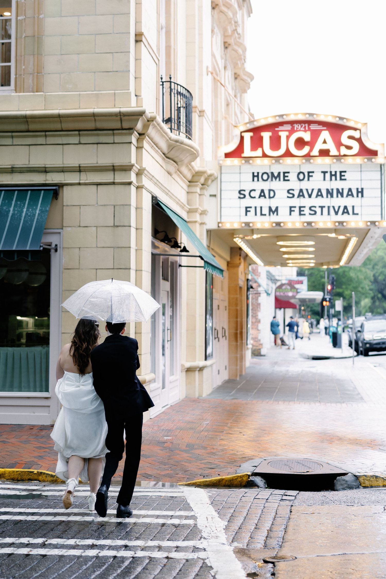 Couple in wedding attire walks under umbrella past Lucas Theatre, home of SCAD Savannah Film Festival, on a rainy day.