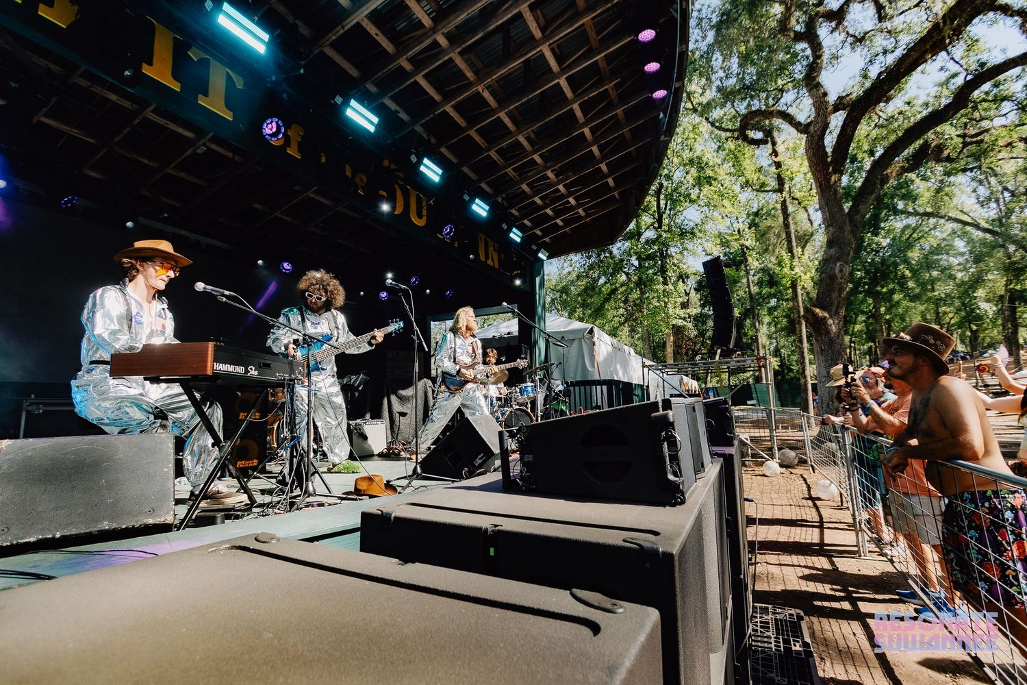 Musicians perform on an outdoor festival stage while attendees watch from a tree-shaded viewing area during daytime.