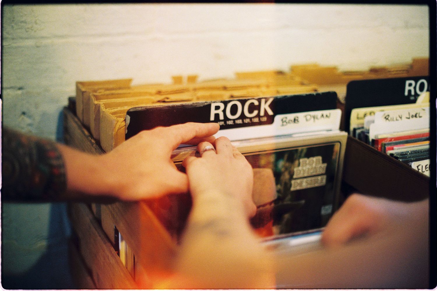 Hands flipping through a crate of vinyl records at a record store, with a divider labeled Rock visible in the bin.