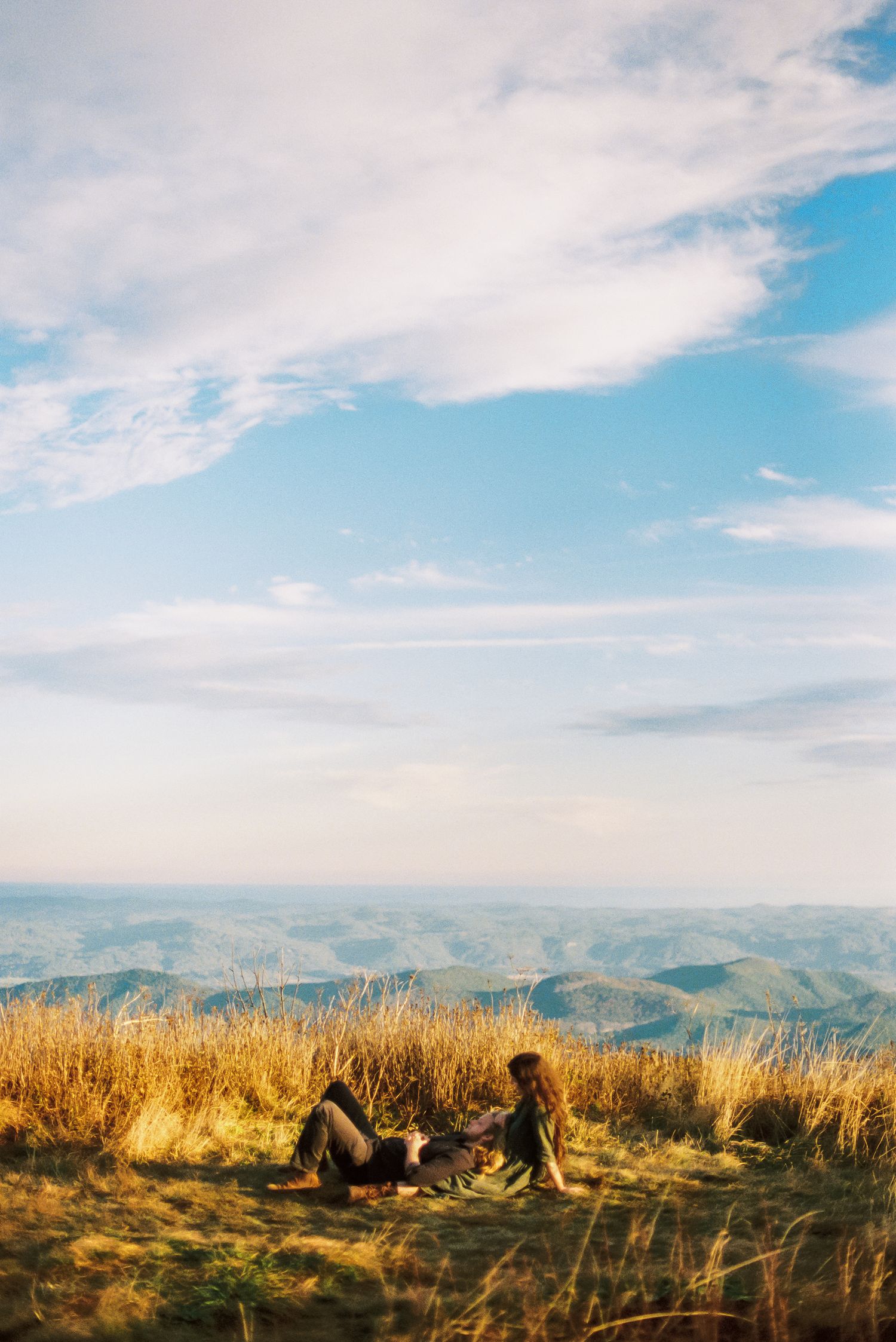 Two people relax in golden grass on a mountain summit, gazing at layered blue ridges under a wide sky of soft clouds.