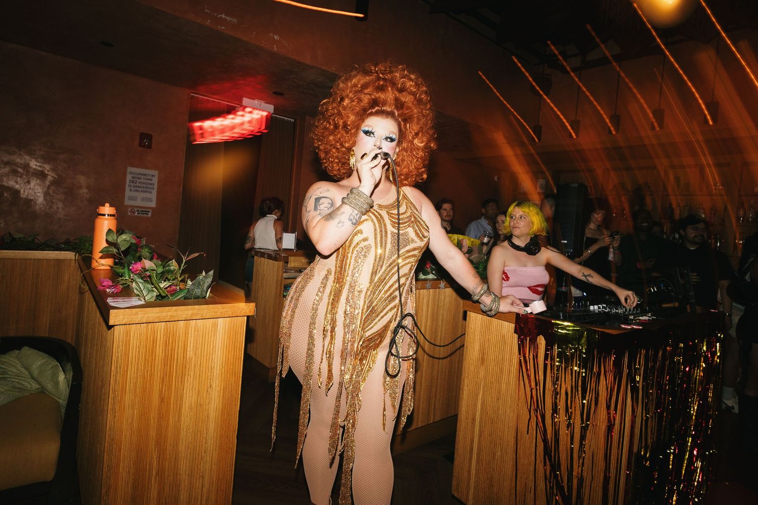 Drag queen performer in gold fringe dress and red curly wig singing on mic at a dimly lit nightclub event.