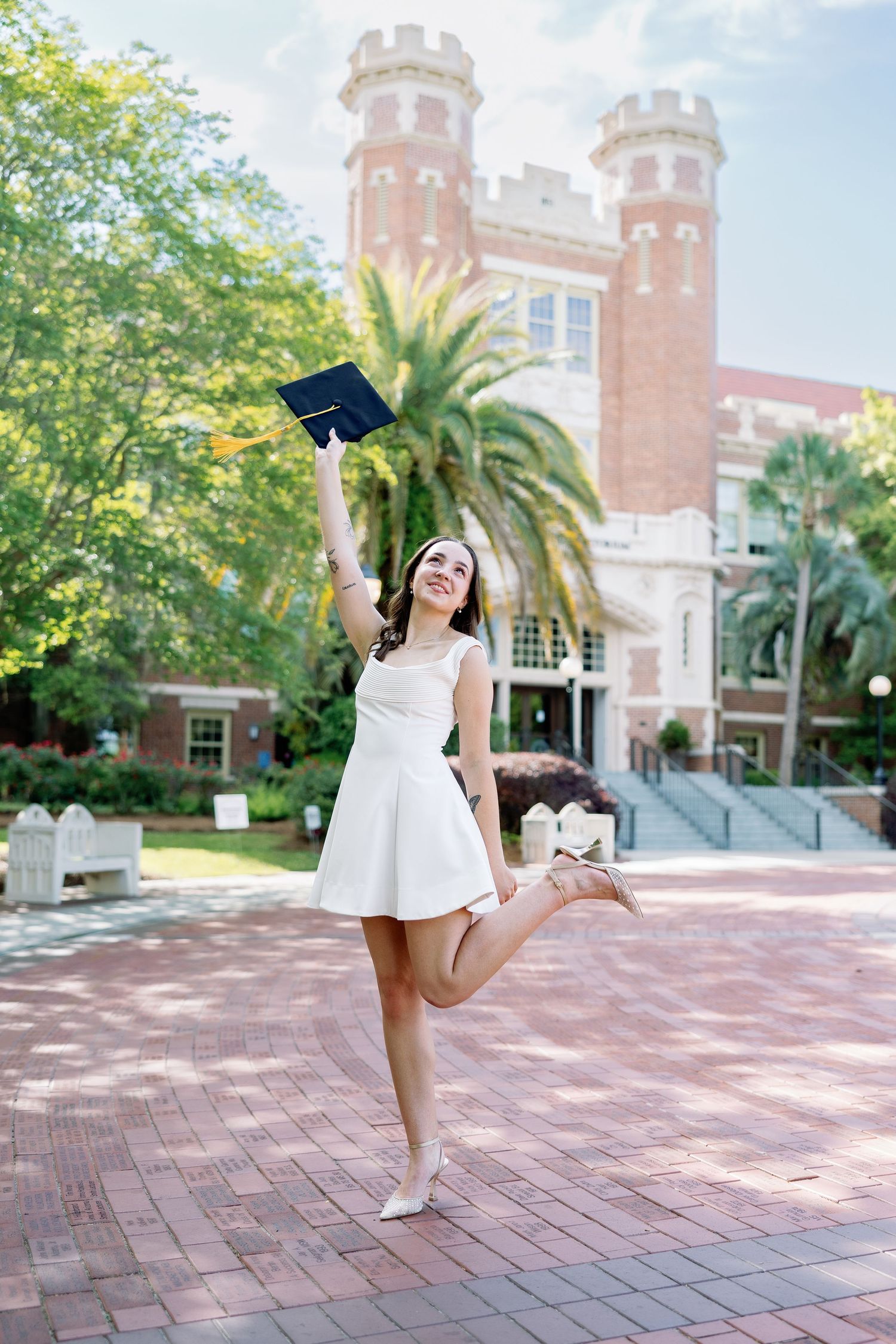 Young woman in white dress celebrating graduation by raising cap in front of brick university building with palm trees.